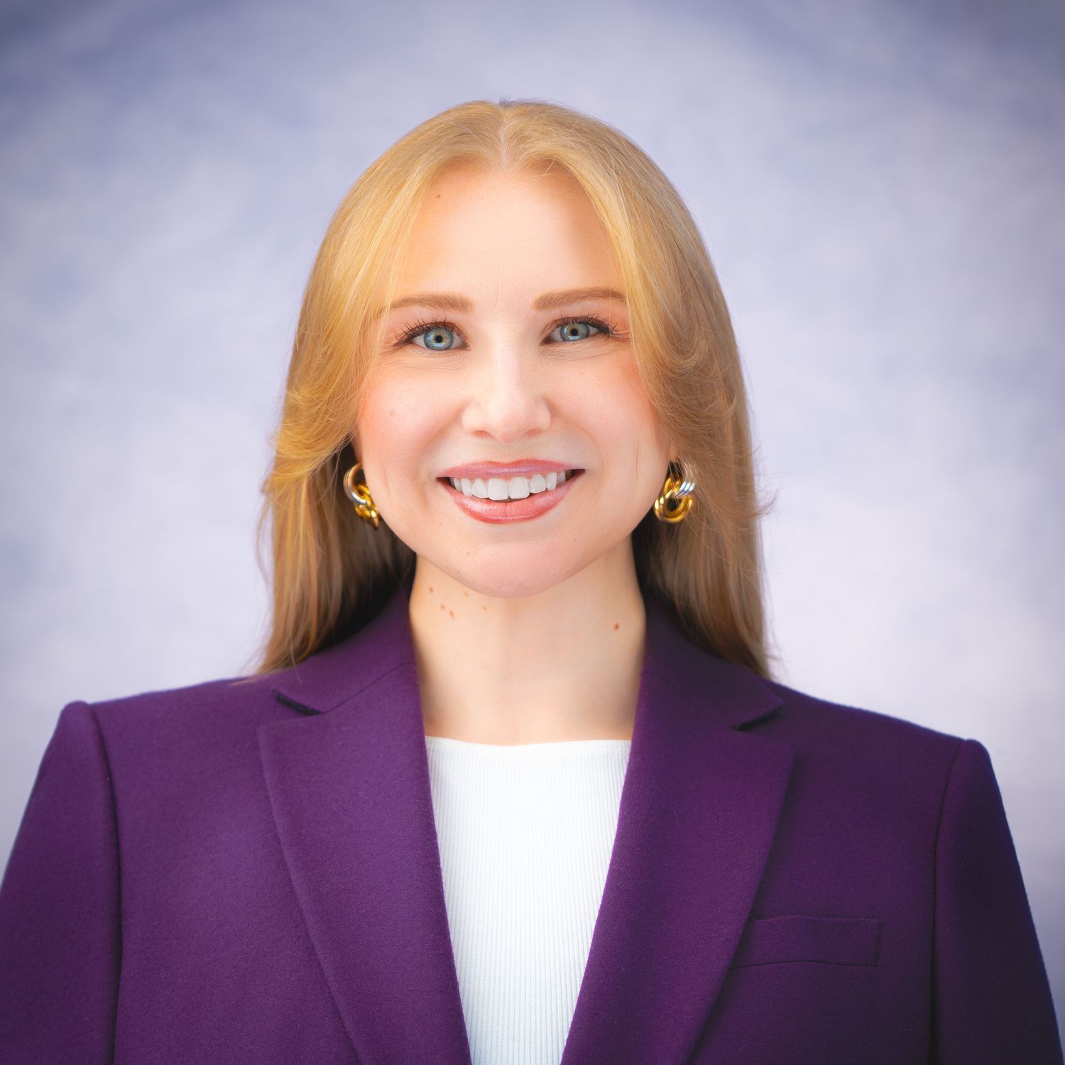 Professional headshot of a person in a purple blazer and white top against a soft gray background.