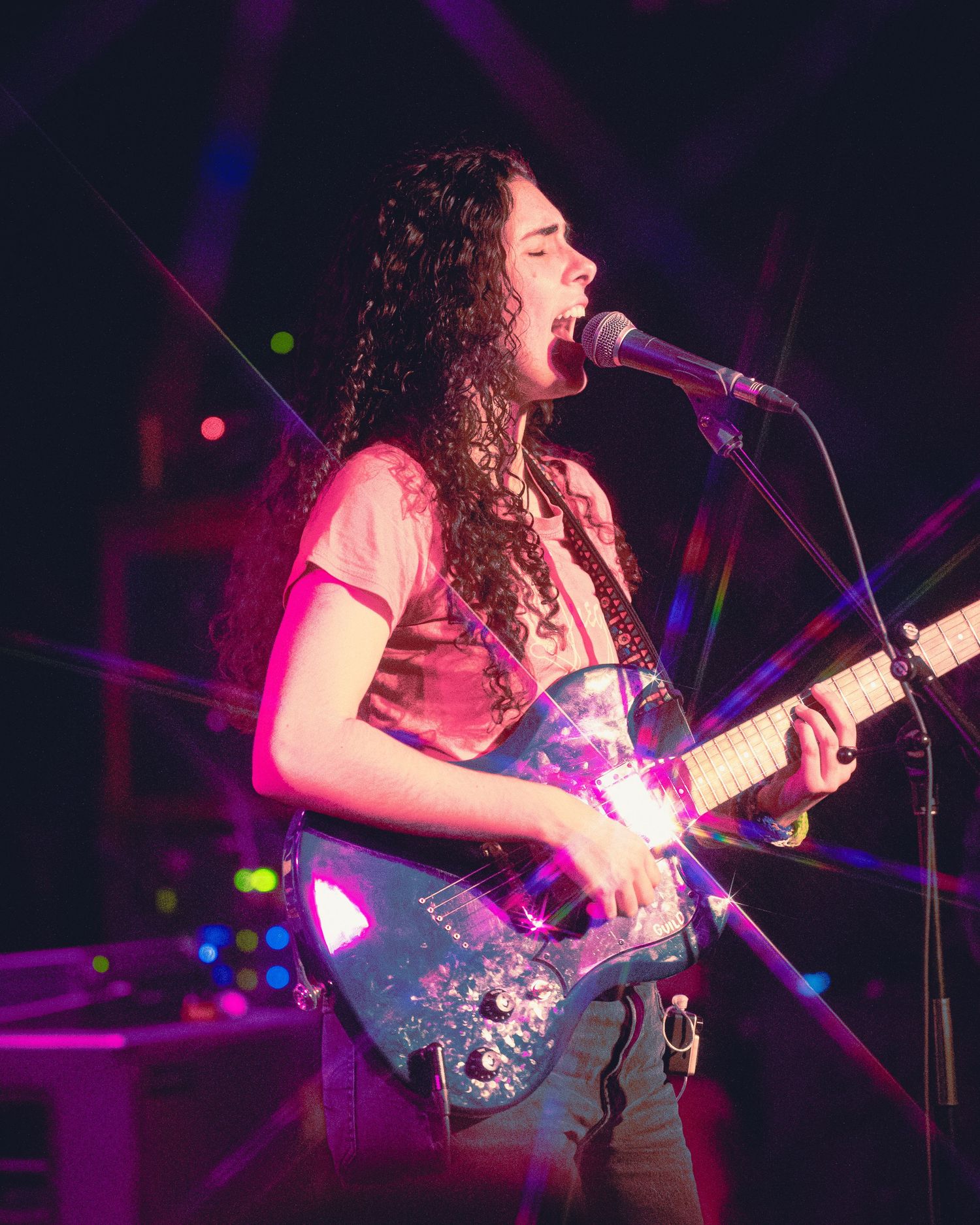 A musician performs on stage with a guitar under dramatic purple and pink lighting during a live concert performance.