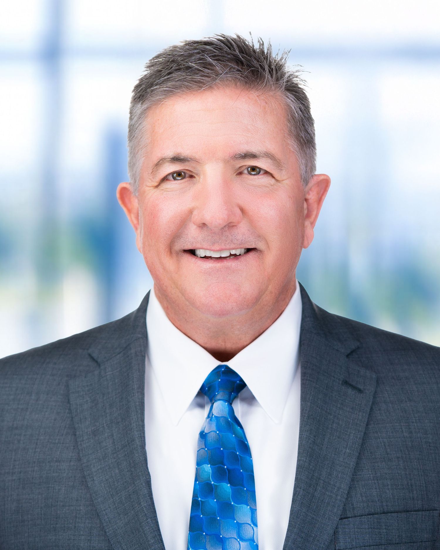 Business headshot of professional wearing charcoal suit and blue patterned tie against bright background.