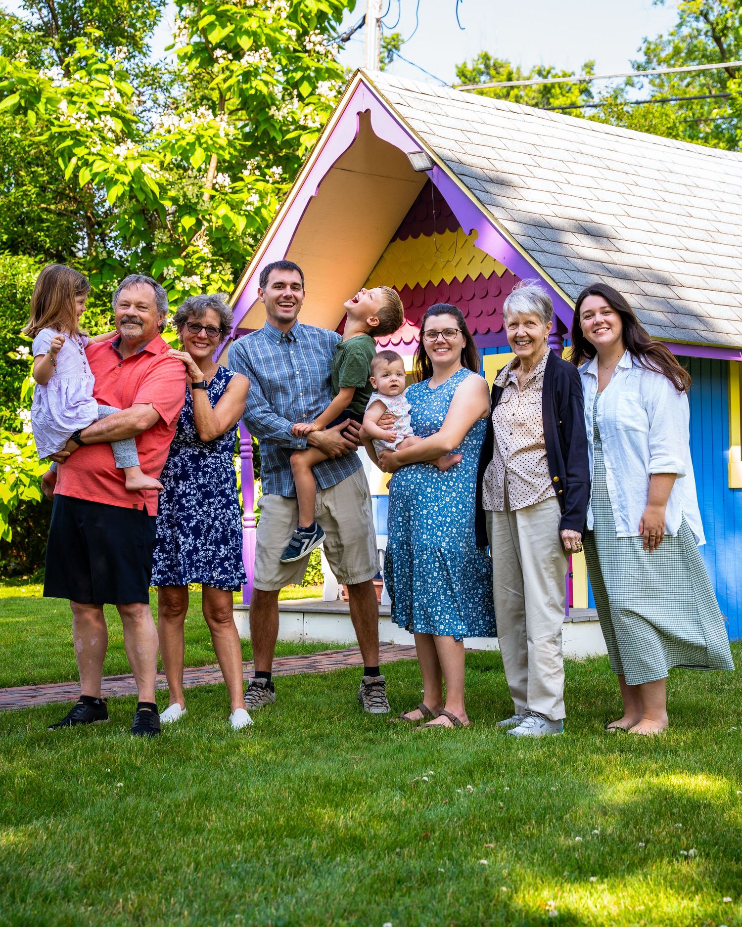 A group stands together smiling on a lawn in front of a whimsical purple and white playhouse on a sunny summer day.