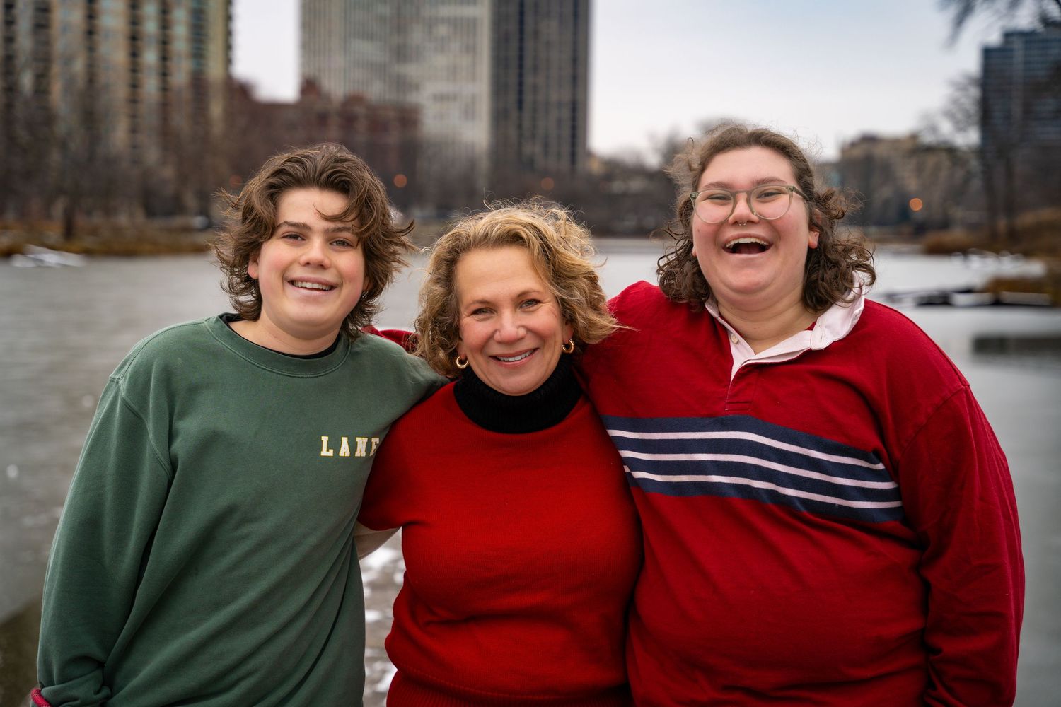 Three people wearing cozy sweaters smile together by a waterfront with city buildings in the background.