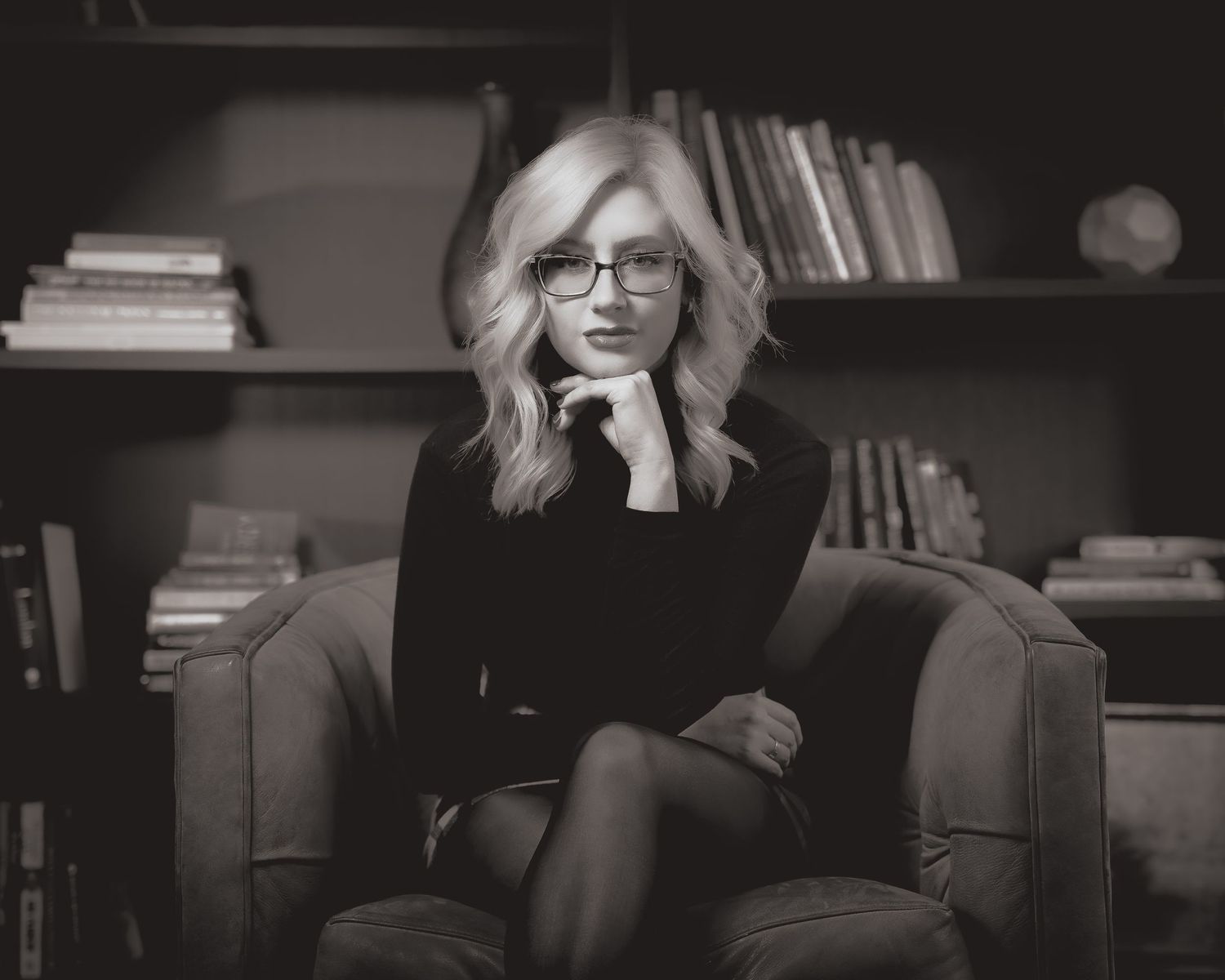 Black and white portrait of someone wearing glasses seated in an armchair with bookshelves in the background.