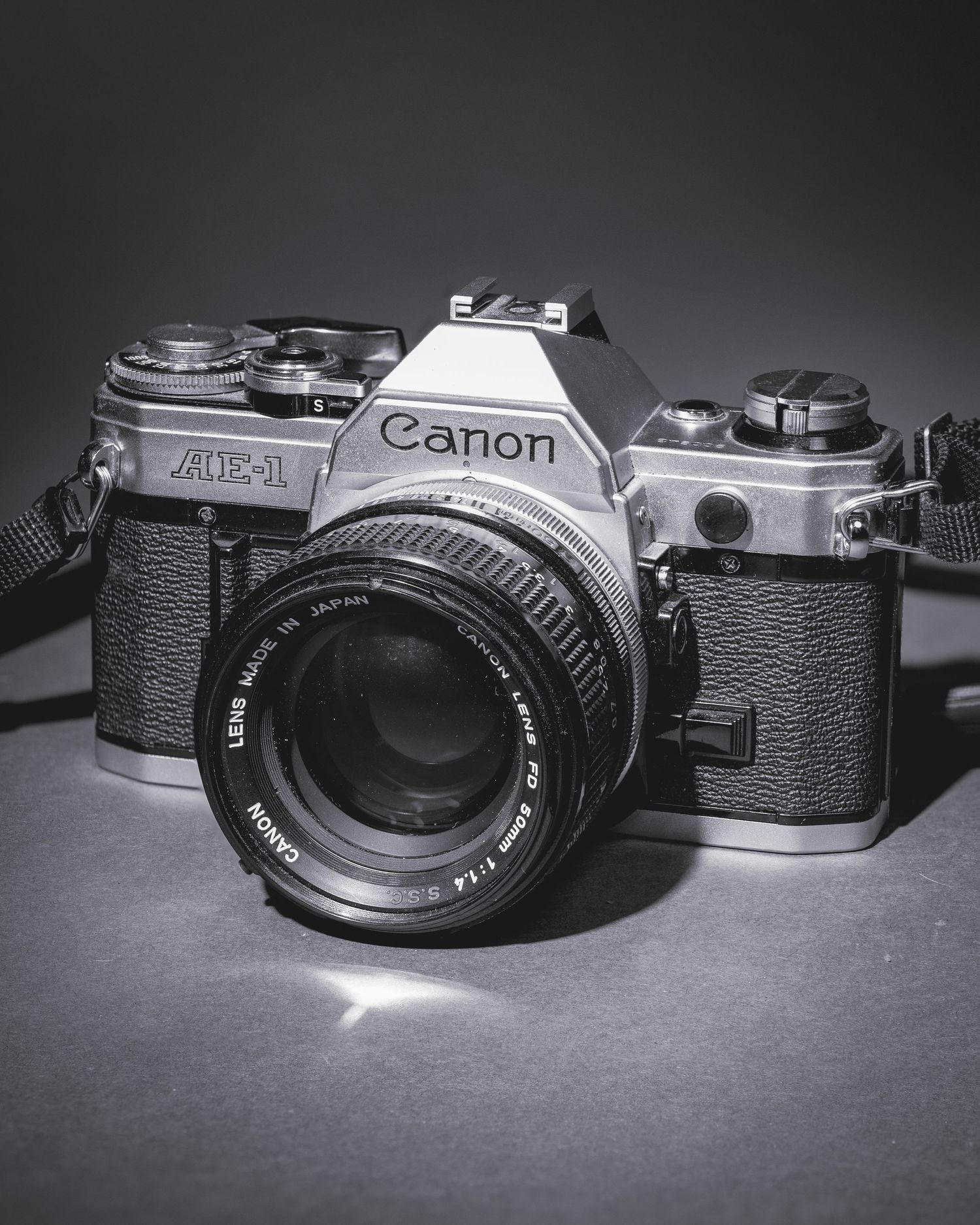 A vintage Canon AE-1 film camera with standard lens displayed in black and white against a dark background.