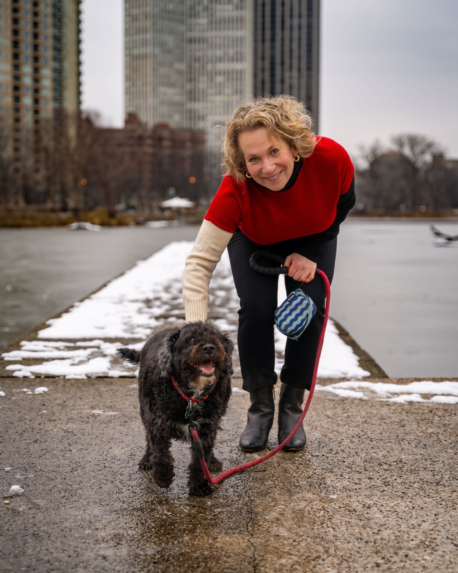 A person in a red sweater walks their shaggy black dog along a snowy path with city buildings in the background.