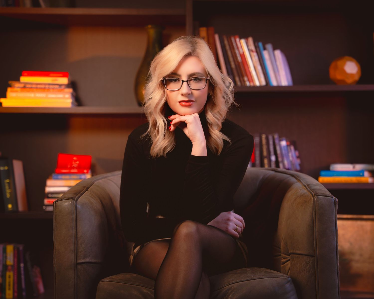 A person in glasses sits in a cozy library setting with bookshelves and warm lighting in the background.