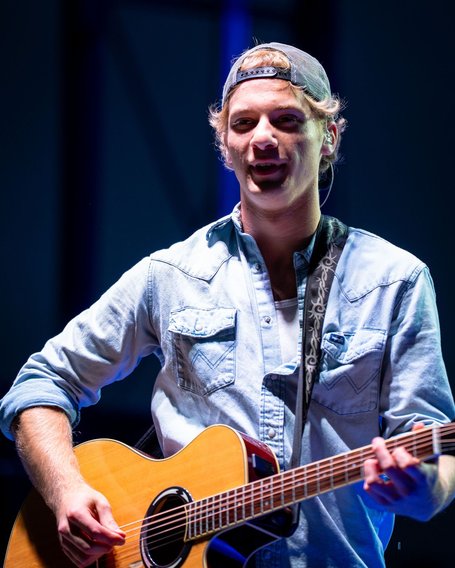 Musician in denim jacket performs on acoustic guitar under stage lighting during live concert.