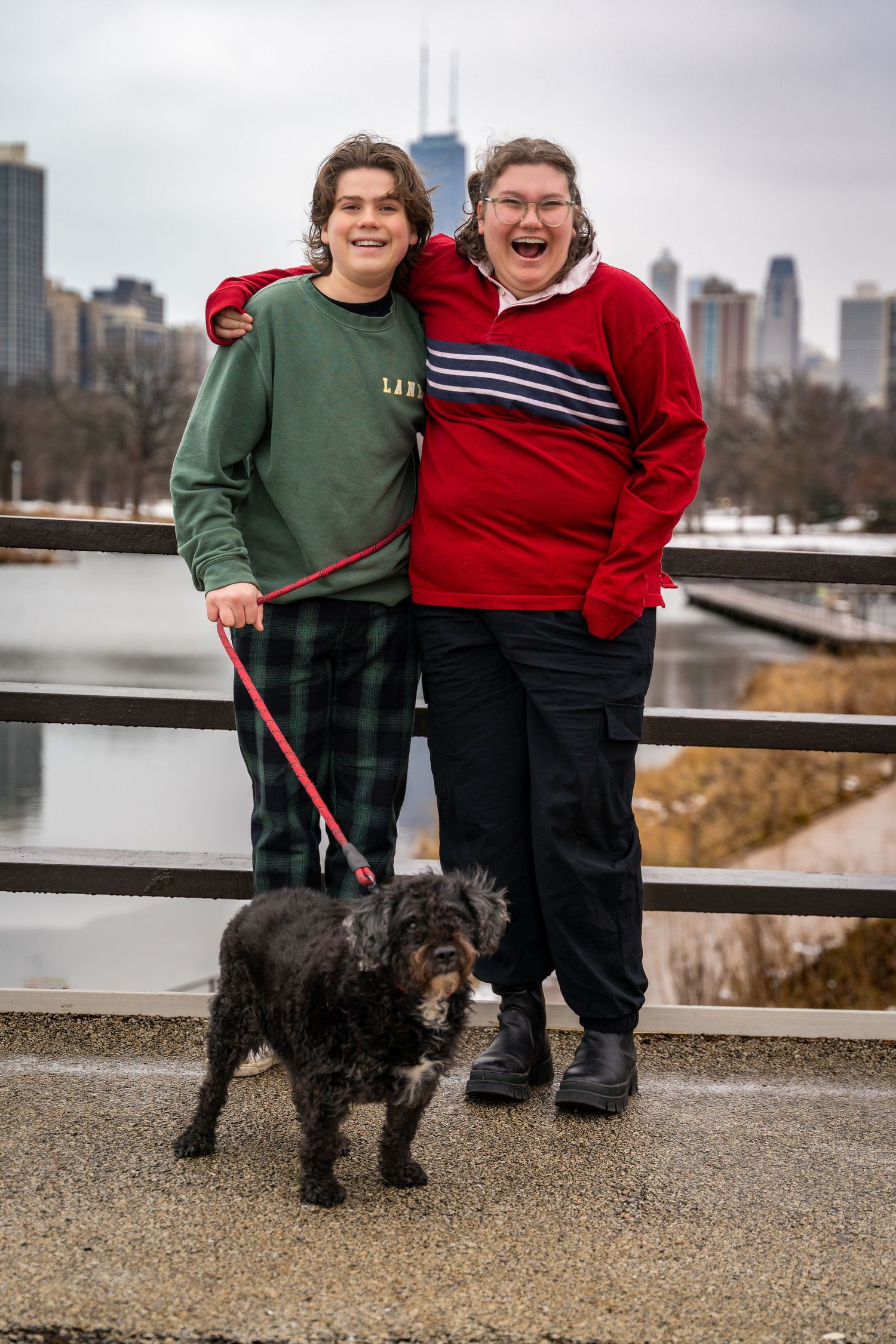Two people in green and red sweaters pose with a black dog on a path overlooking a city skyline.