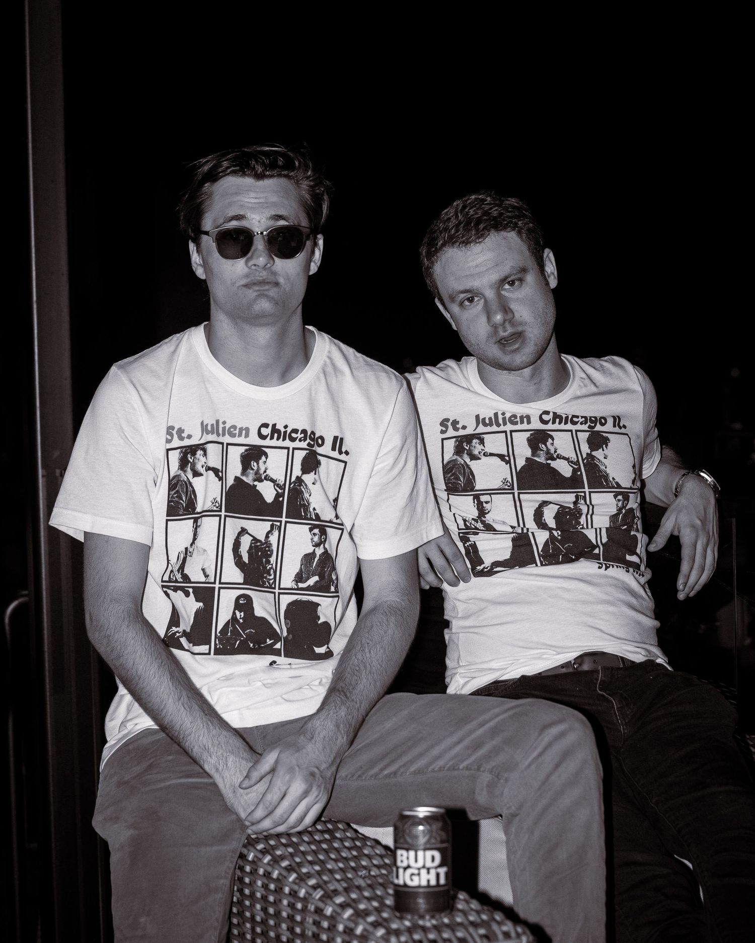 Two band members wearing sunglasses and matching t-shirts sit together.