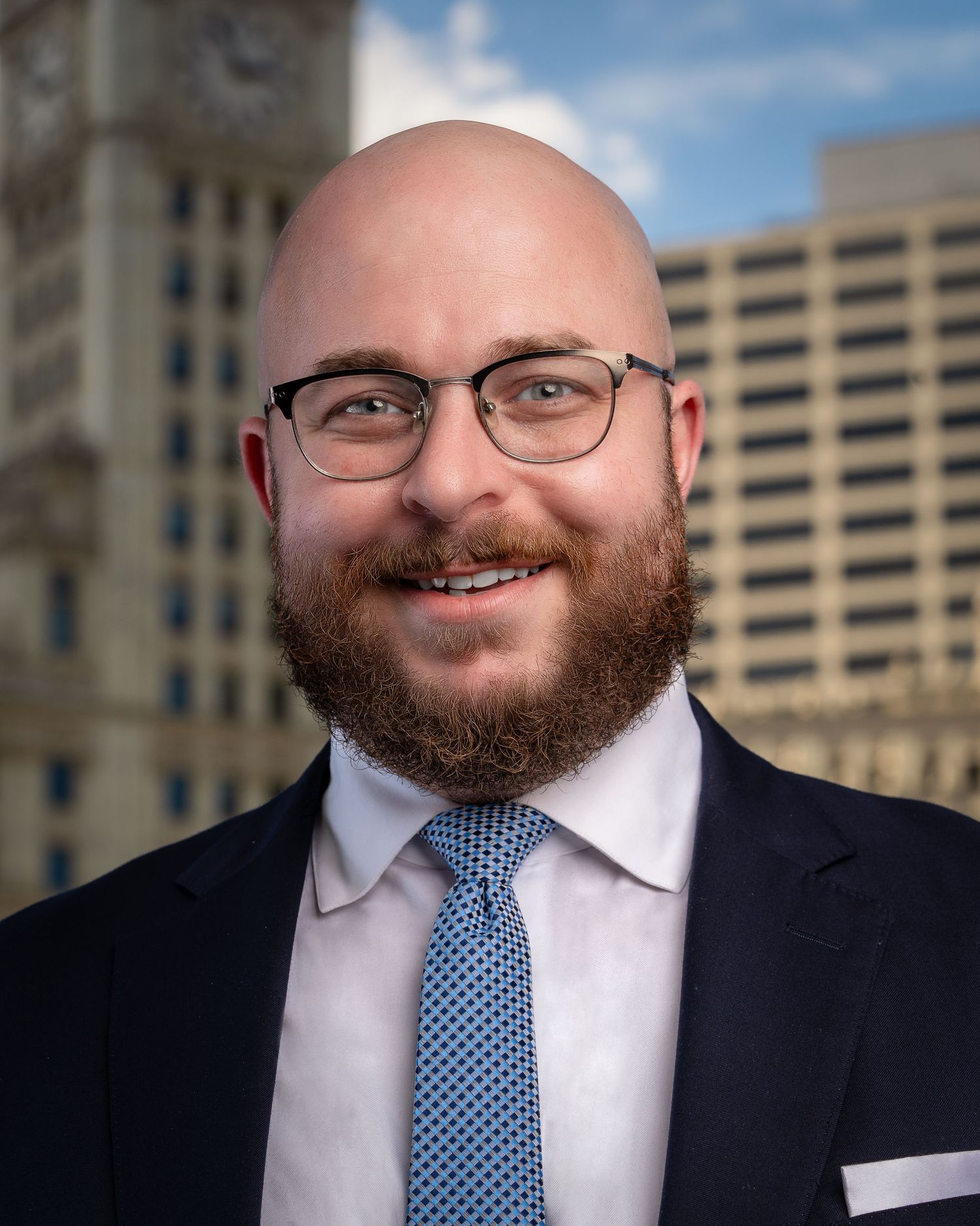 Professional headshot series against urban architecture background wearing navy suit and blue patterned tie.