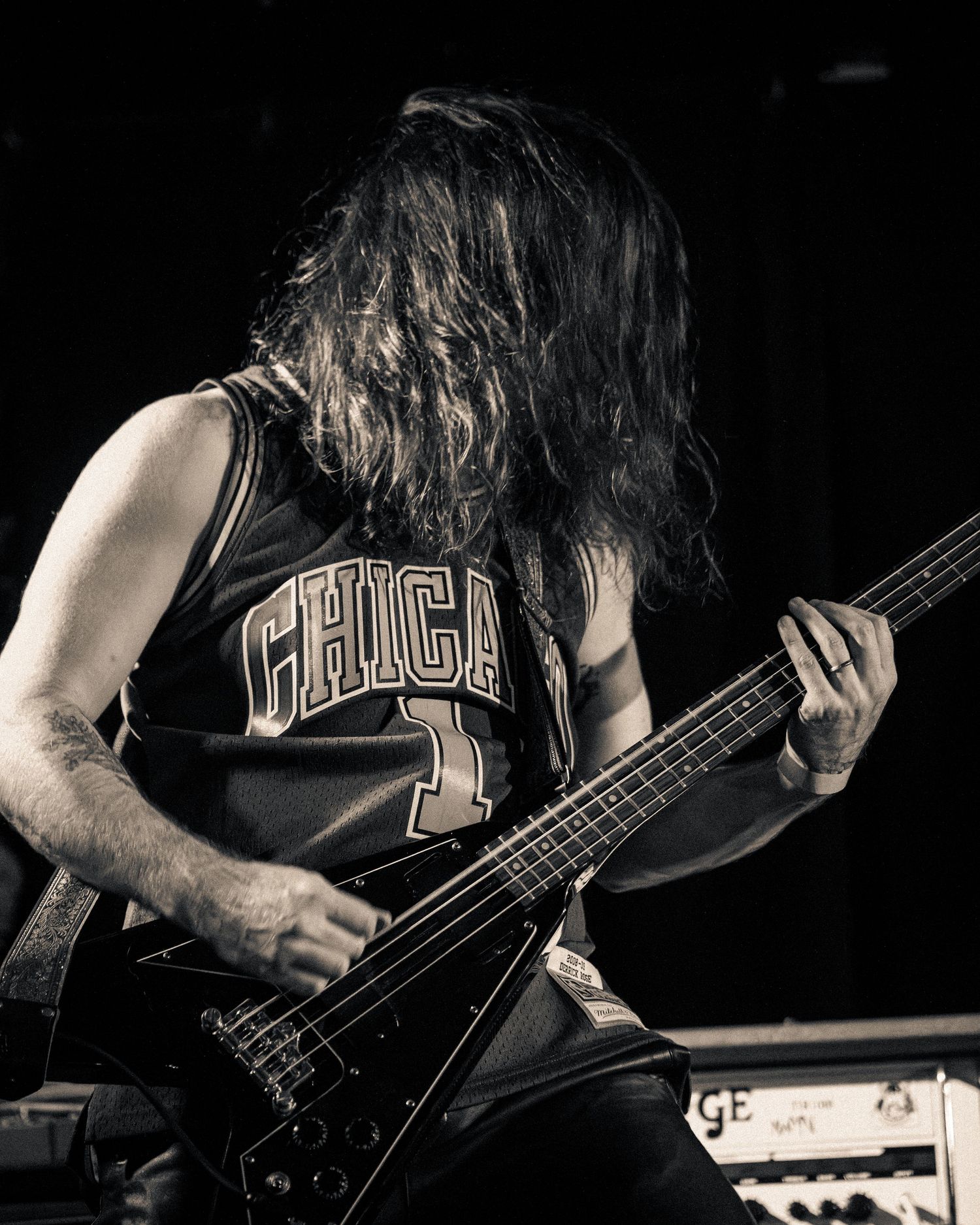 Bass guitarist in dark clothing performs on stage with dramatic side lighting.