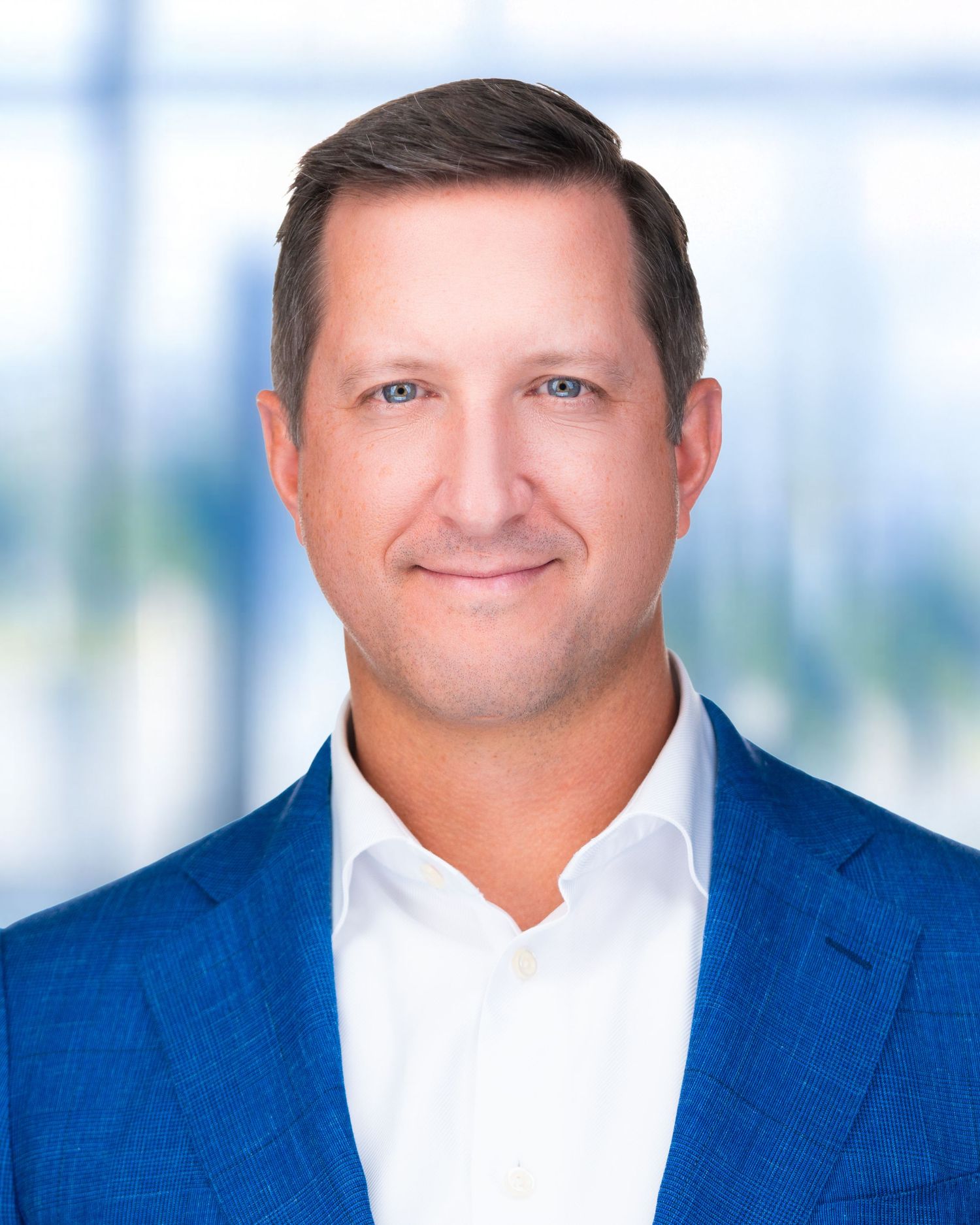 Professional portrait of person in white shirt and navy jacket against modern office backdrop.