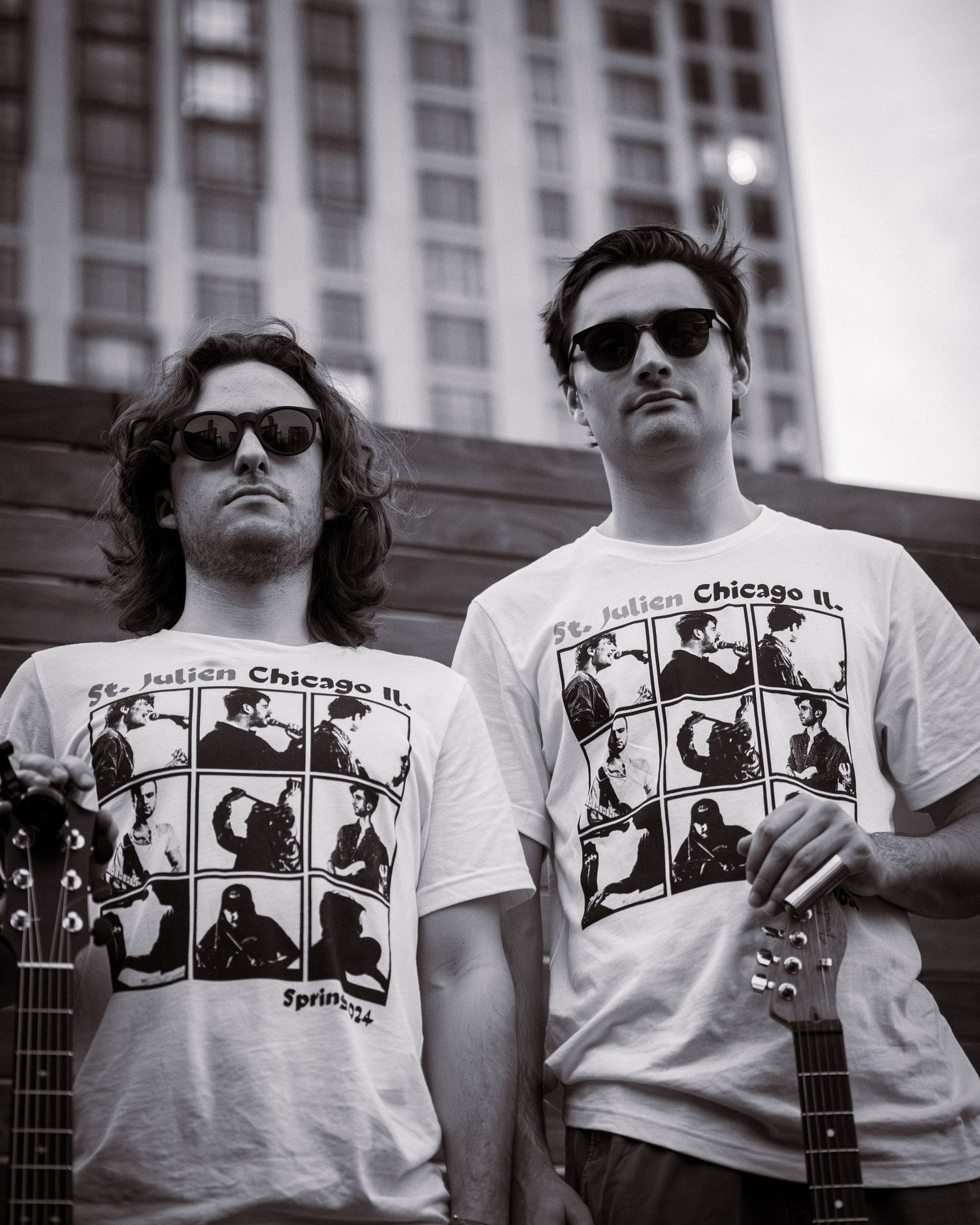 Two musicians with sunglasses wearing matching graphic t-shirts pose with guitars.