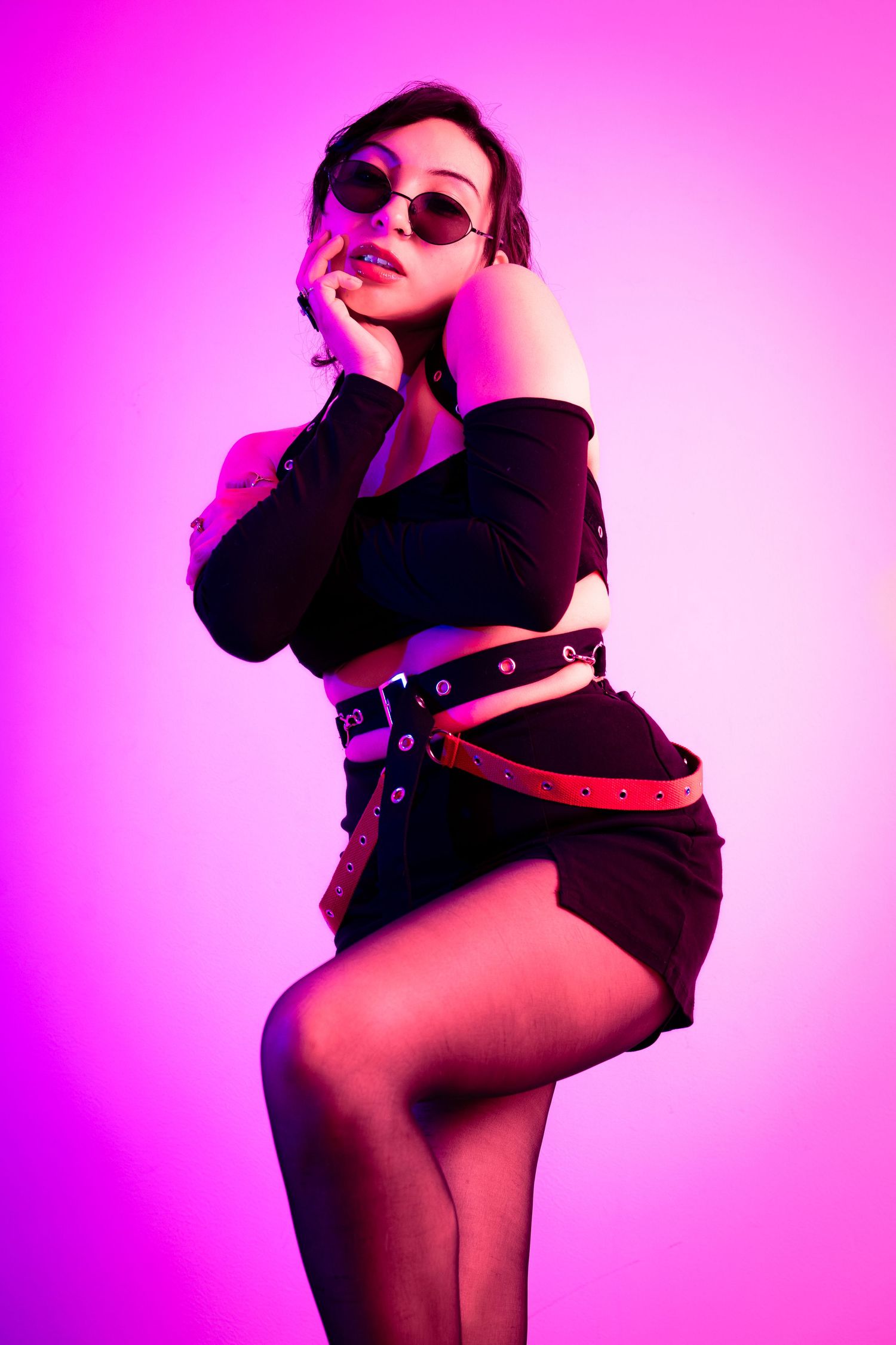 Stylish fashion pose in black crop top and sunglasses against bright pink background.