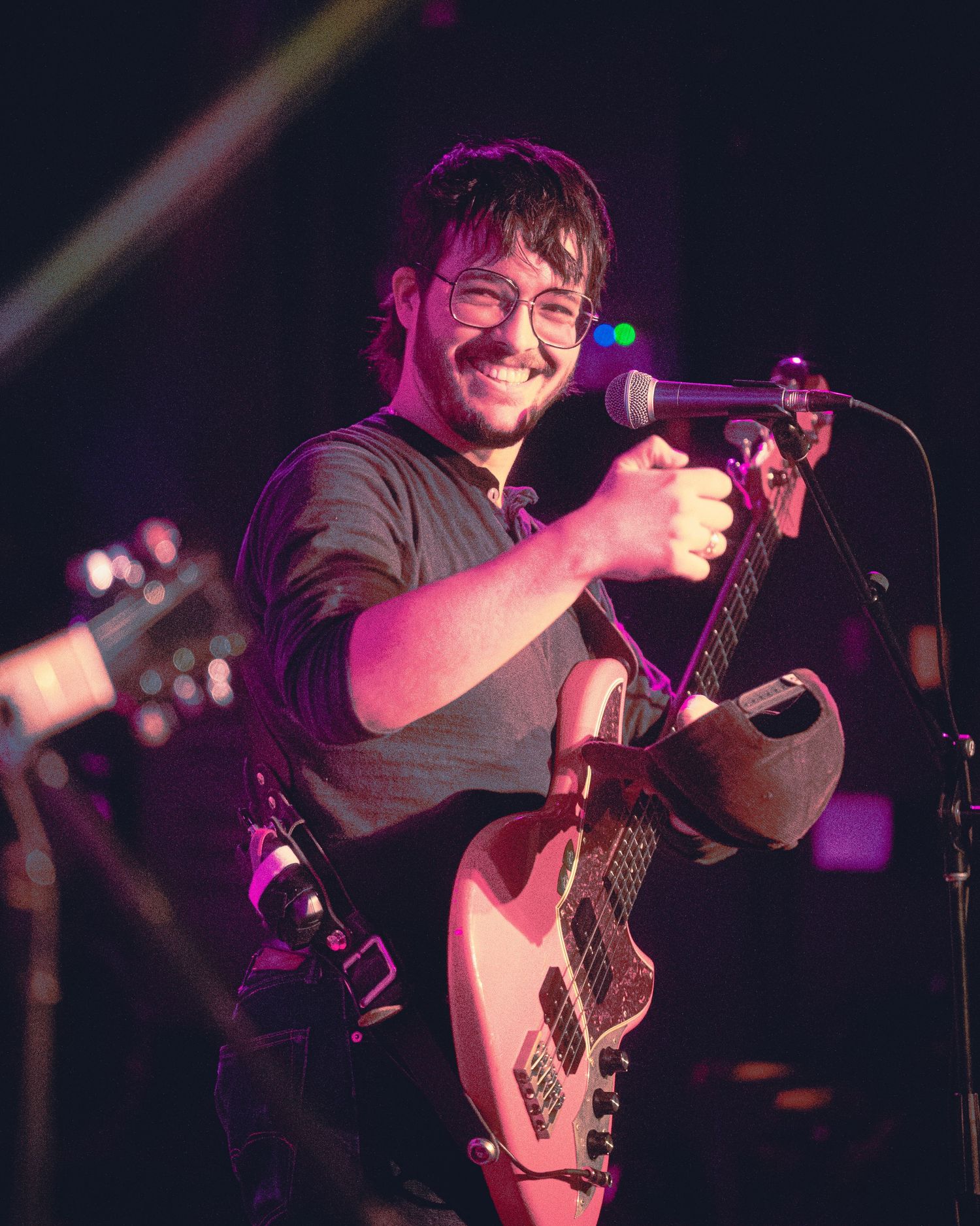 A musician performs on stage with a bass guitar under purple and blue lighting during a live concert performance.