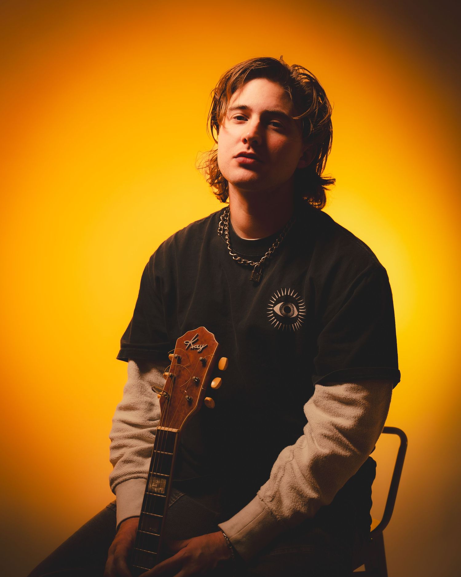 Musician in black clothing sitting with guitar against warm orange backdrop lighting.