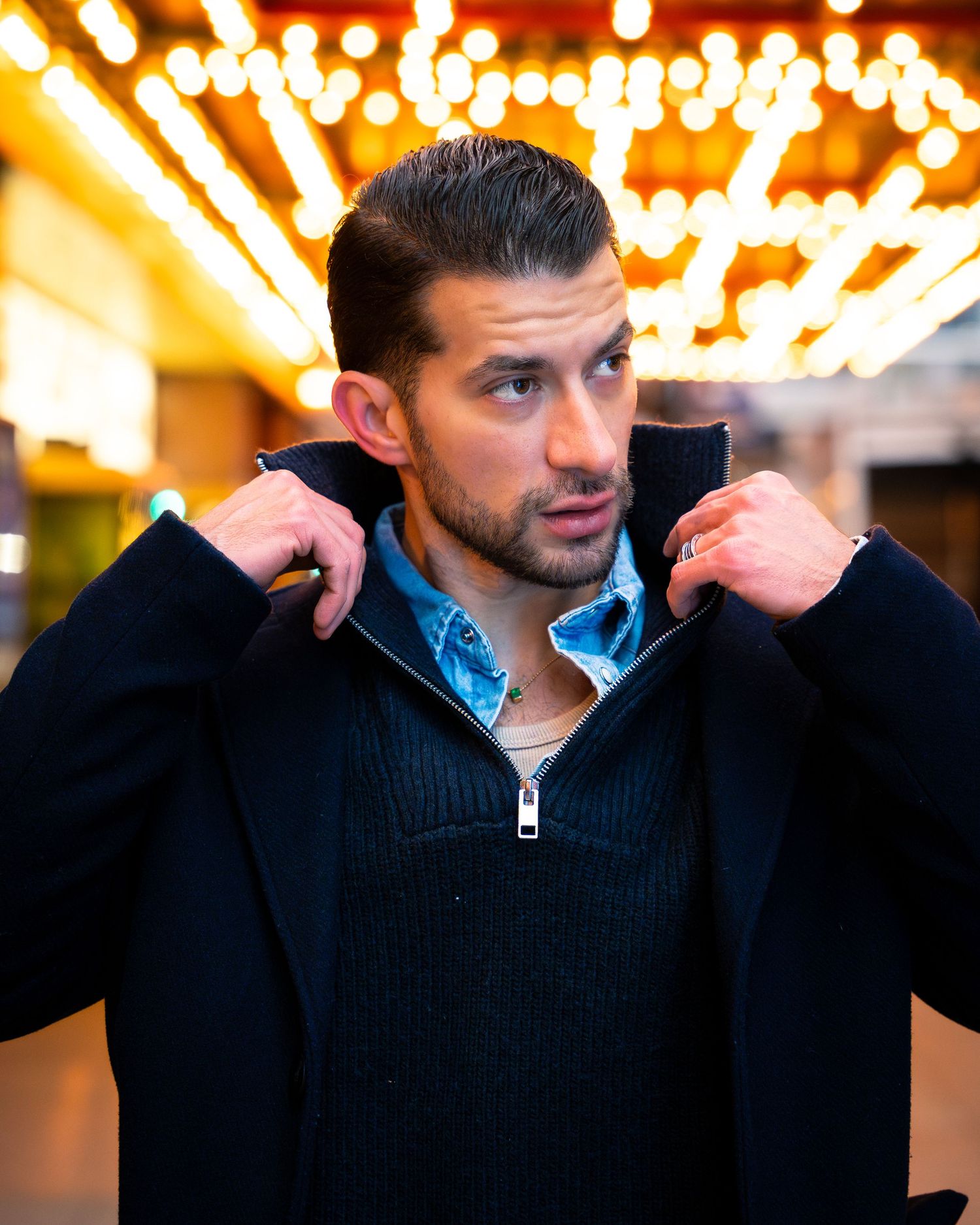 Man in dark sweater poses with festive lights bokeh in background.