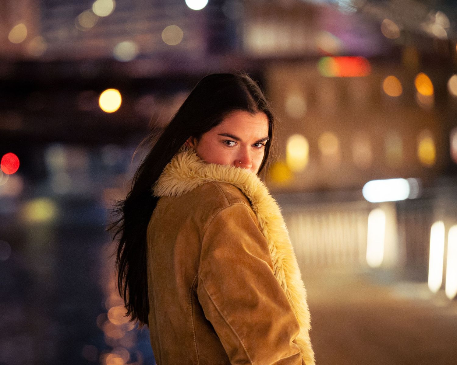 Person in tan fur coat photographed at night with bokeh lights in background.