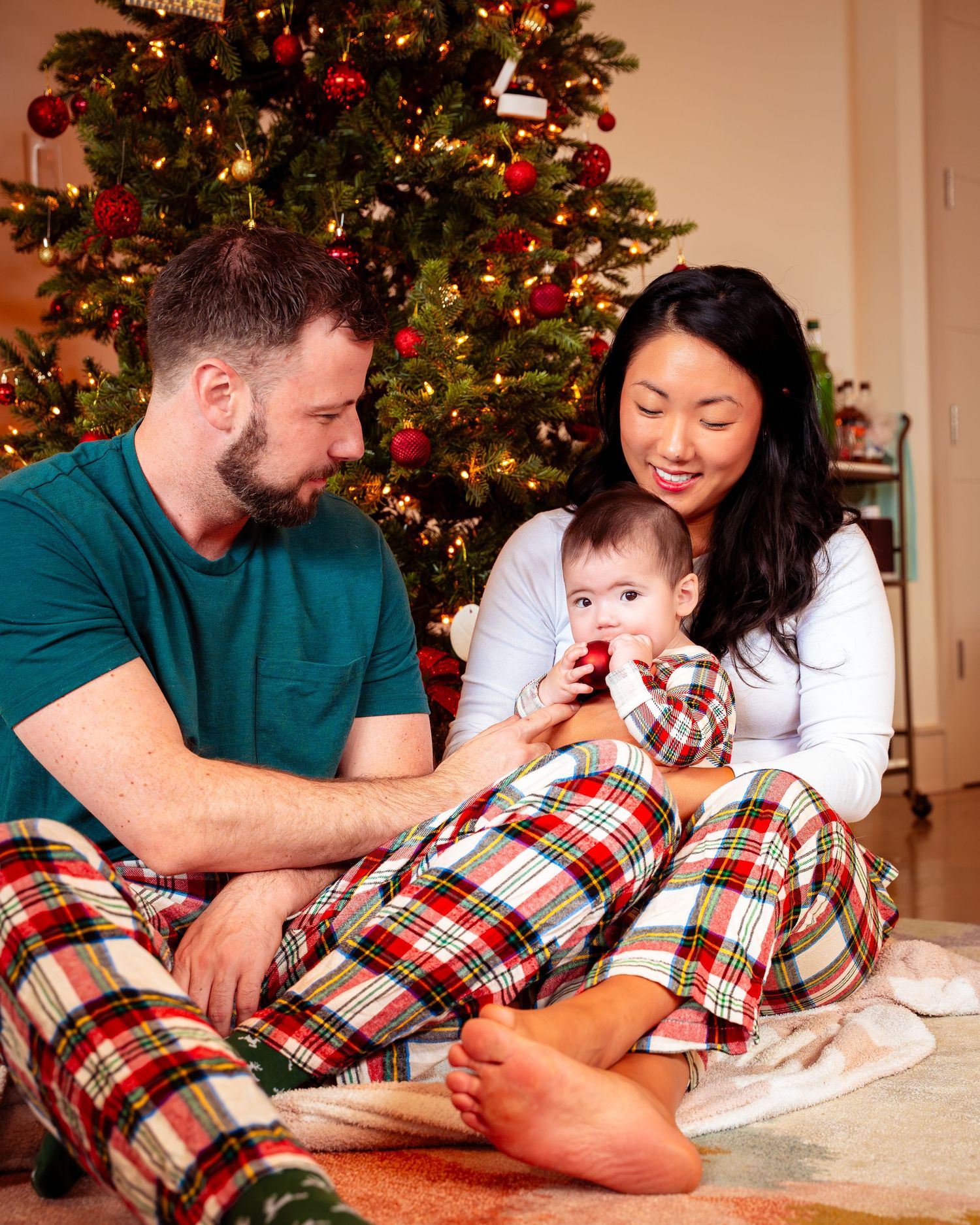 Family wearing matching plaid pajamas lies on floor together near decorated Christmas tree.