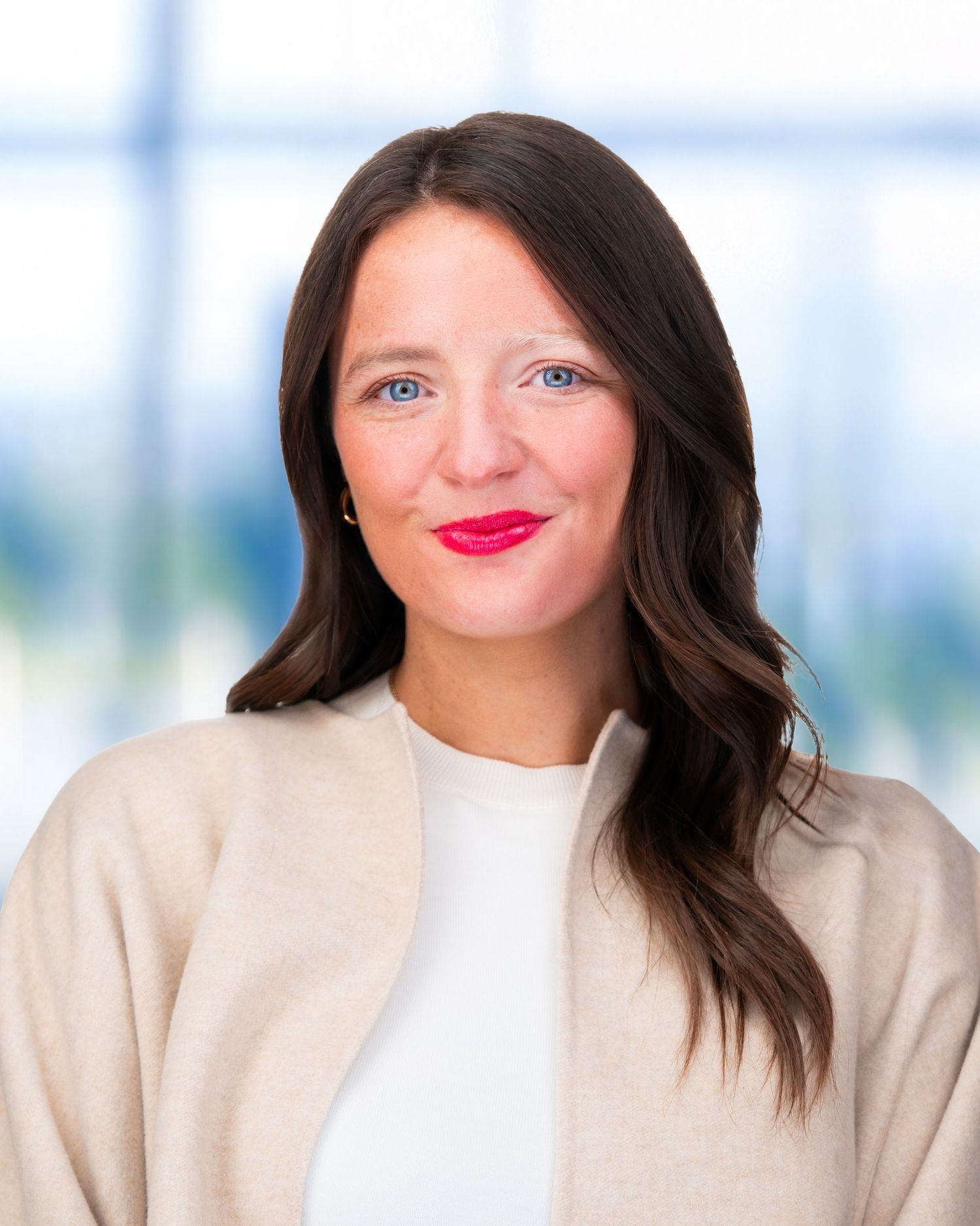 Professional headshot of woman in beige sweater with bright red lipstick against window background.