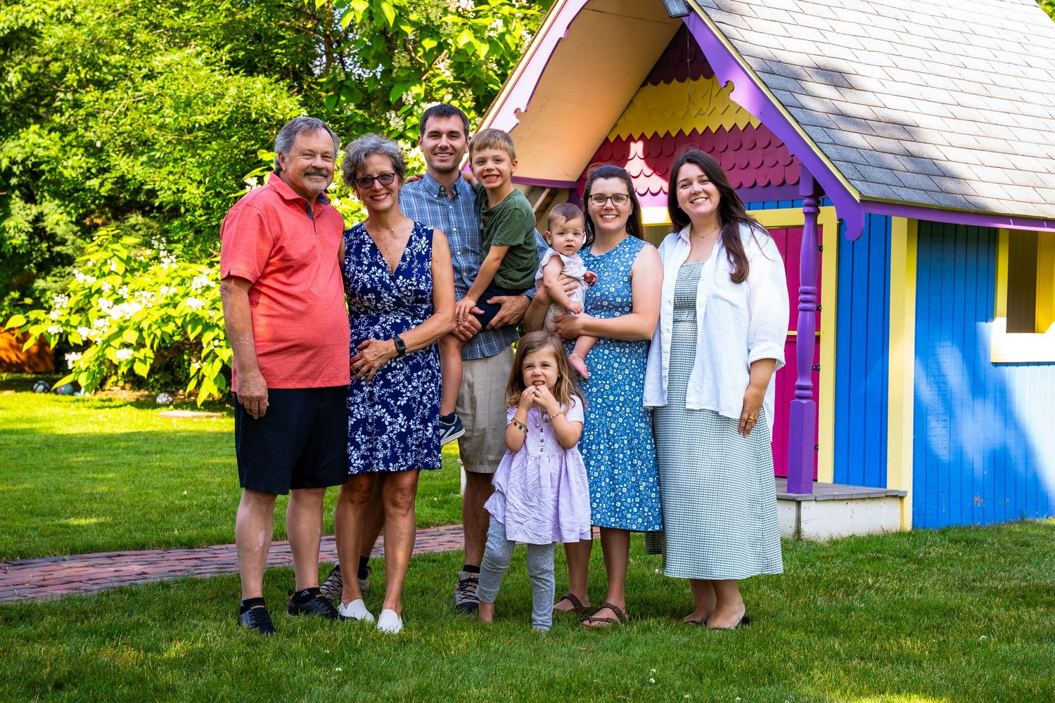 A group of people gather together on a sunny day in front of a colorful playhouse structure in a grassy yard.