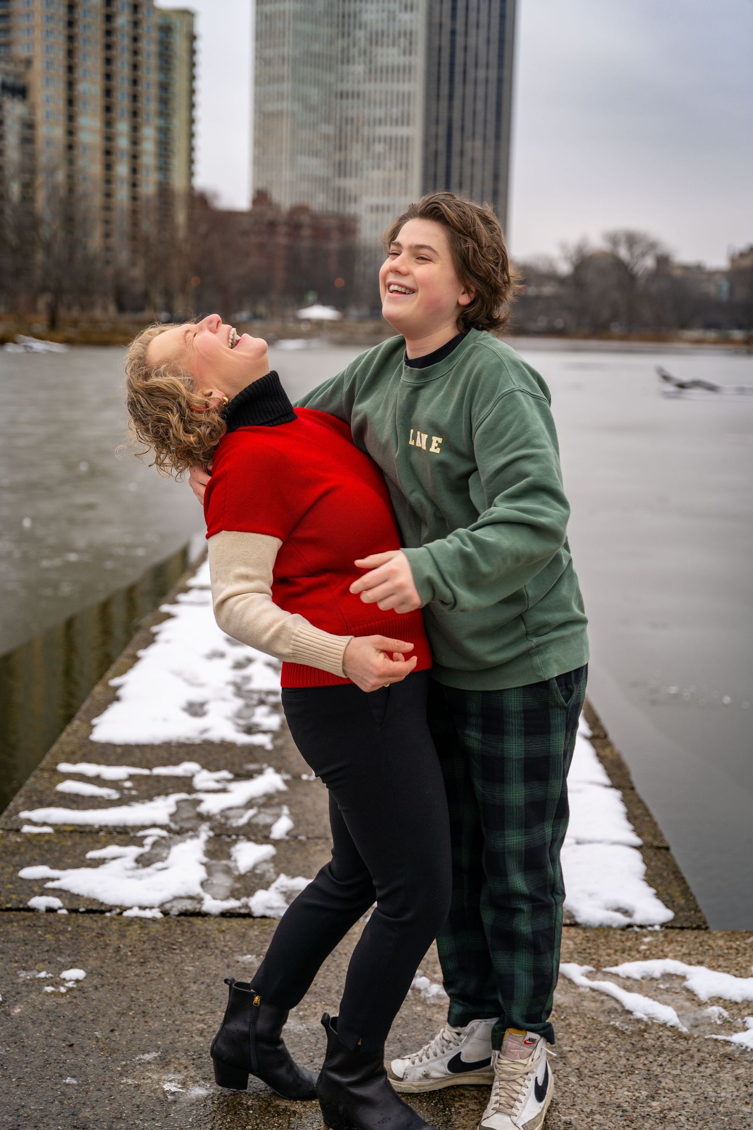 Two people laughing and embracing while standing near a snowy waterfront with city buildings in the background.