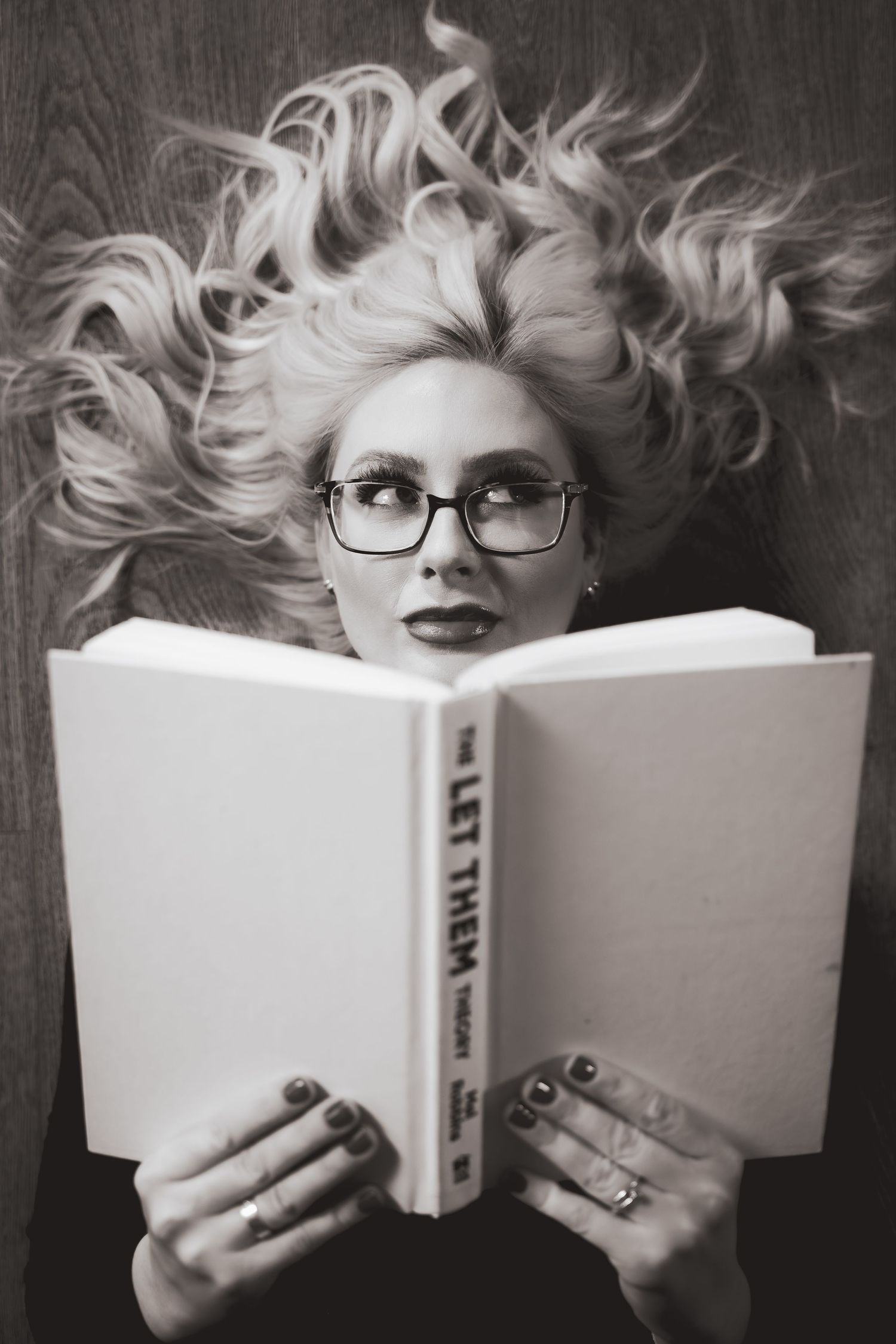 Black and white artistic photo of someone with flowing white hair wearing glasses while reading a book.