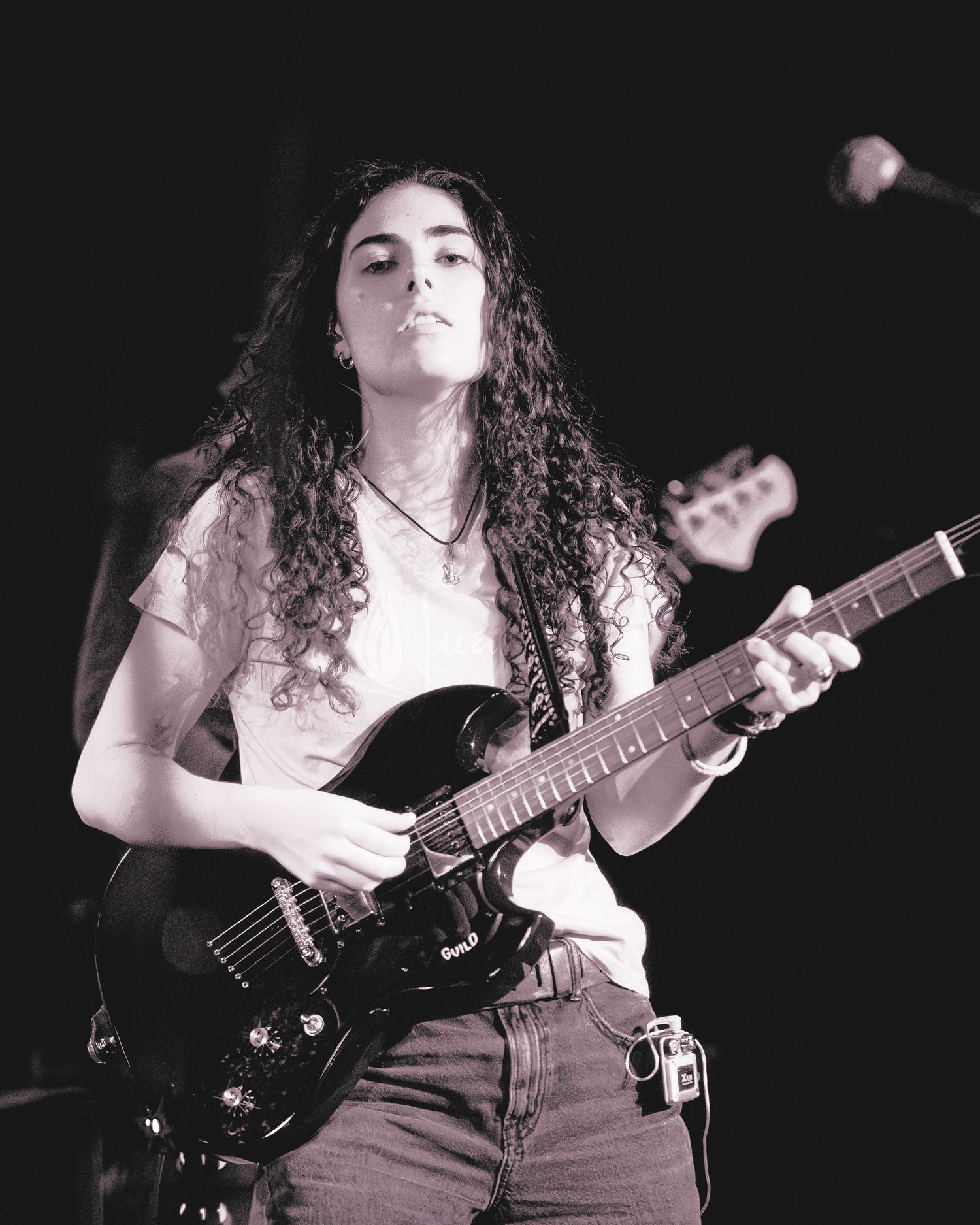 A musician plays bass guitar on a dark stage wearing a white top, captured in dramatic black and white lighting.