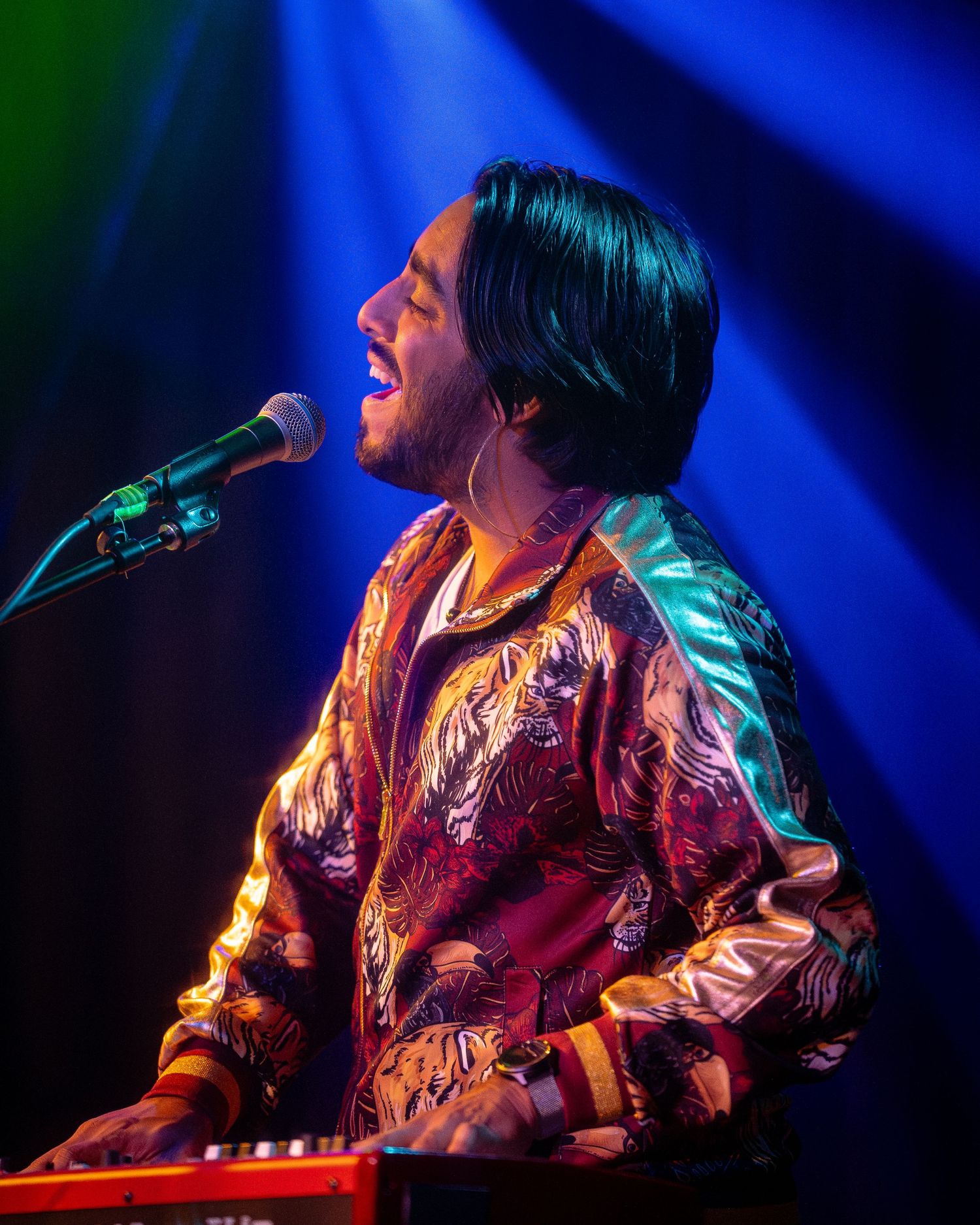 Performer in shiny jacket singing emotionally under blue and green stage lights.