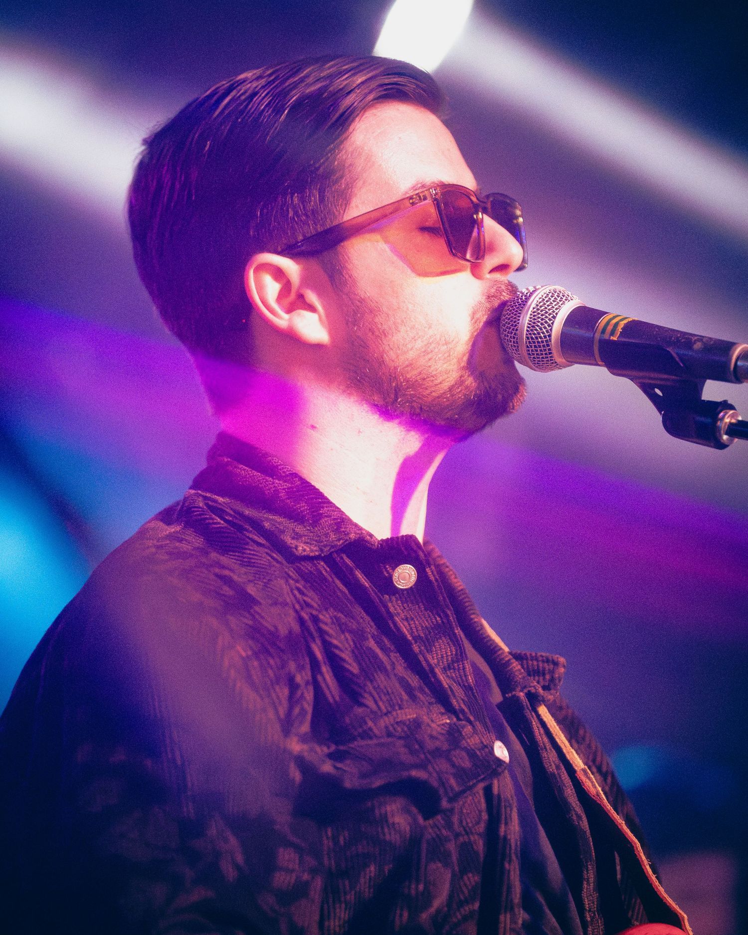 Musician performing on stage with dramatic purple lighting and sunglasses singing into a microphone.
