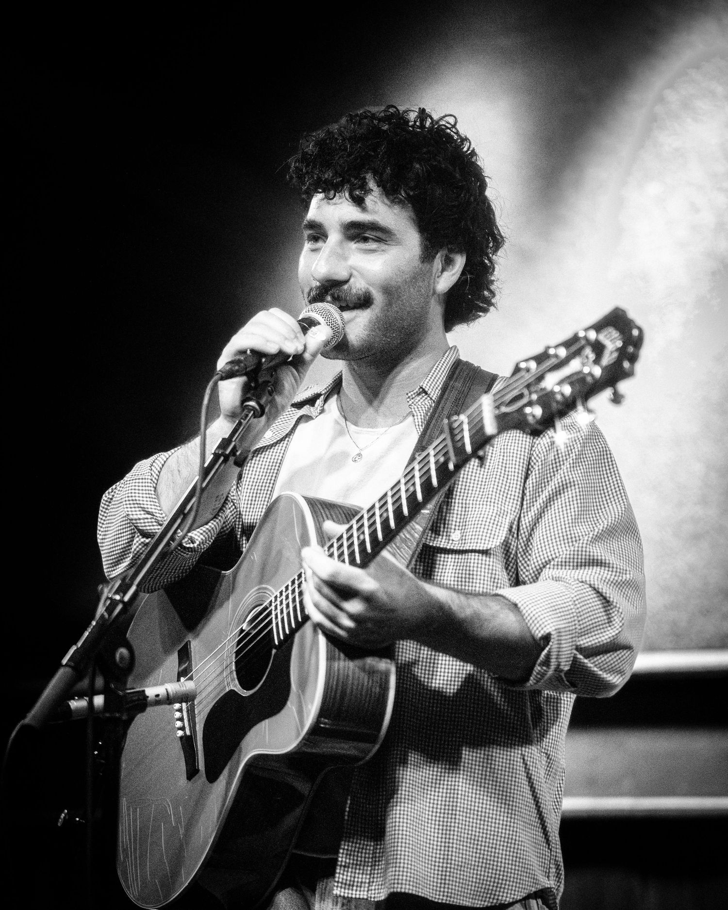 Black and white performance shot showing musician playing acoustic guitar on illuminated stage.