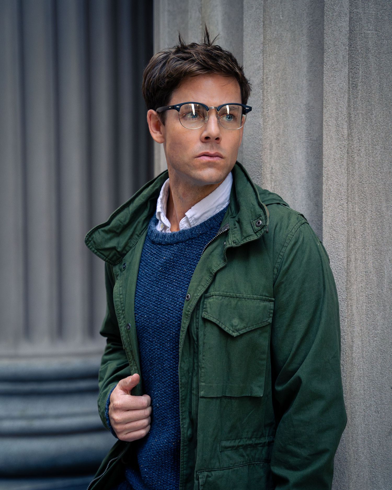 Professional fashion photo of a green utility jacket paired with navy sweater and glasses against a stone column background.