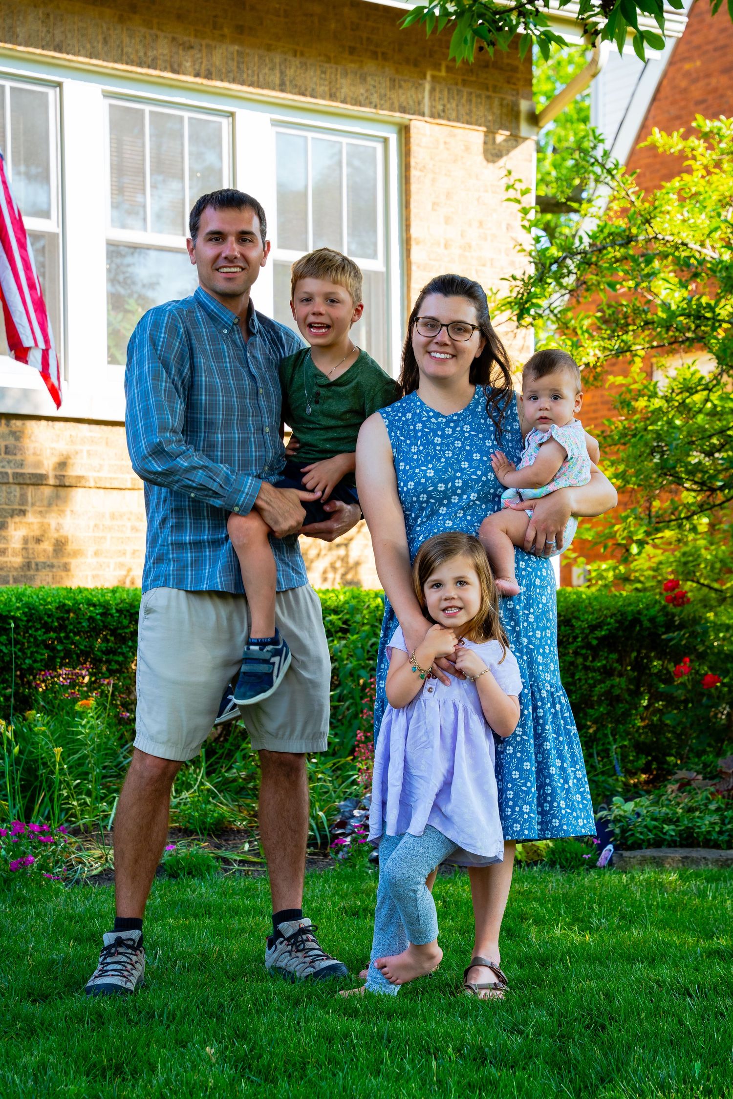 A family poses together on their front lawn with flowering bushes and greenery in the background.