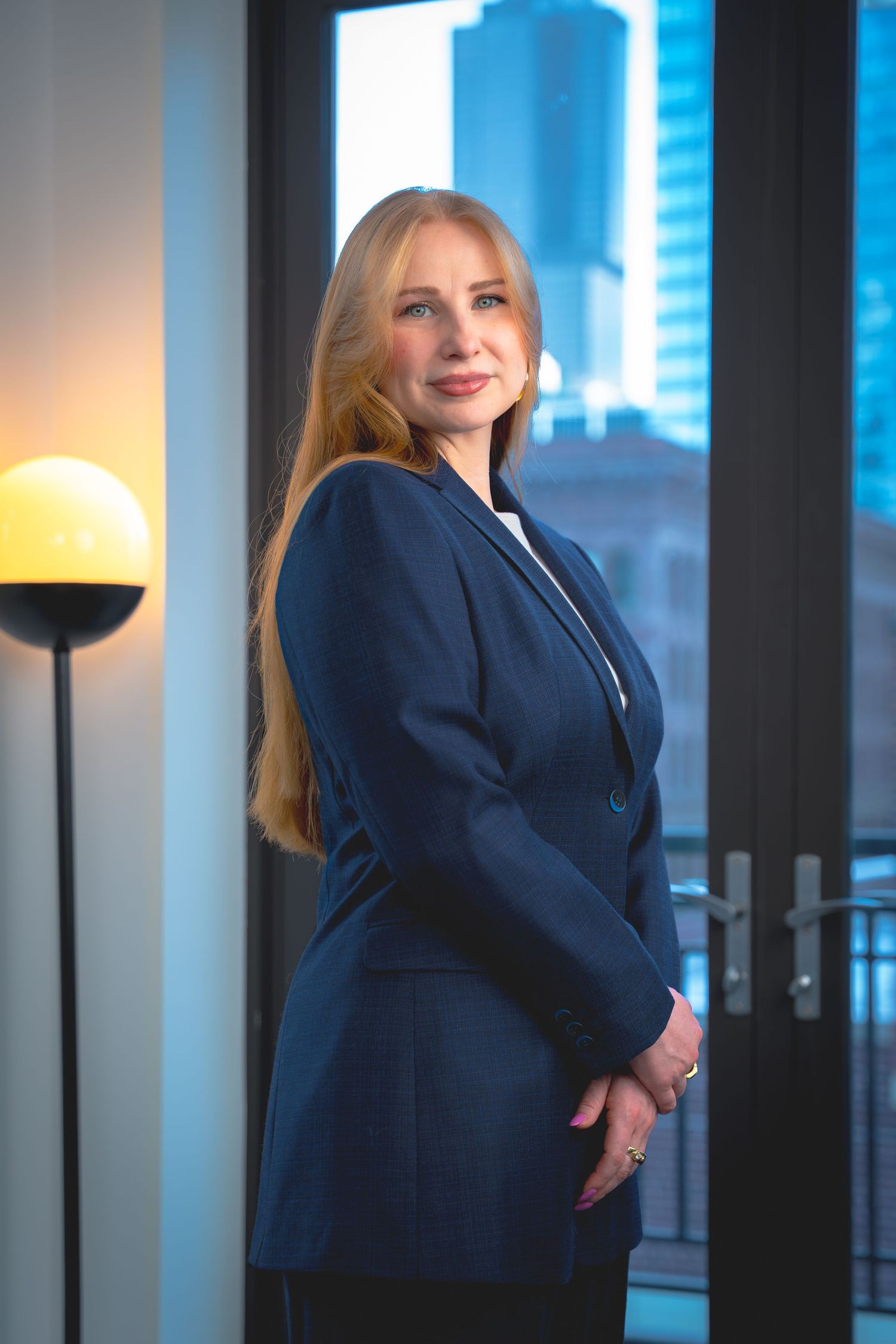 Professional portrait in a navy blazer against a modern office backdrop with city views.