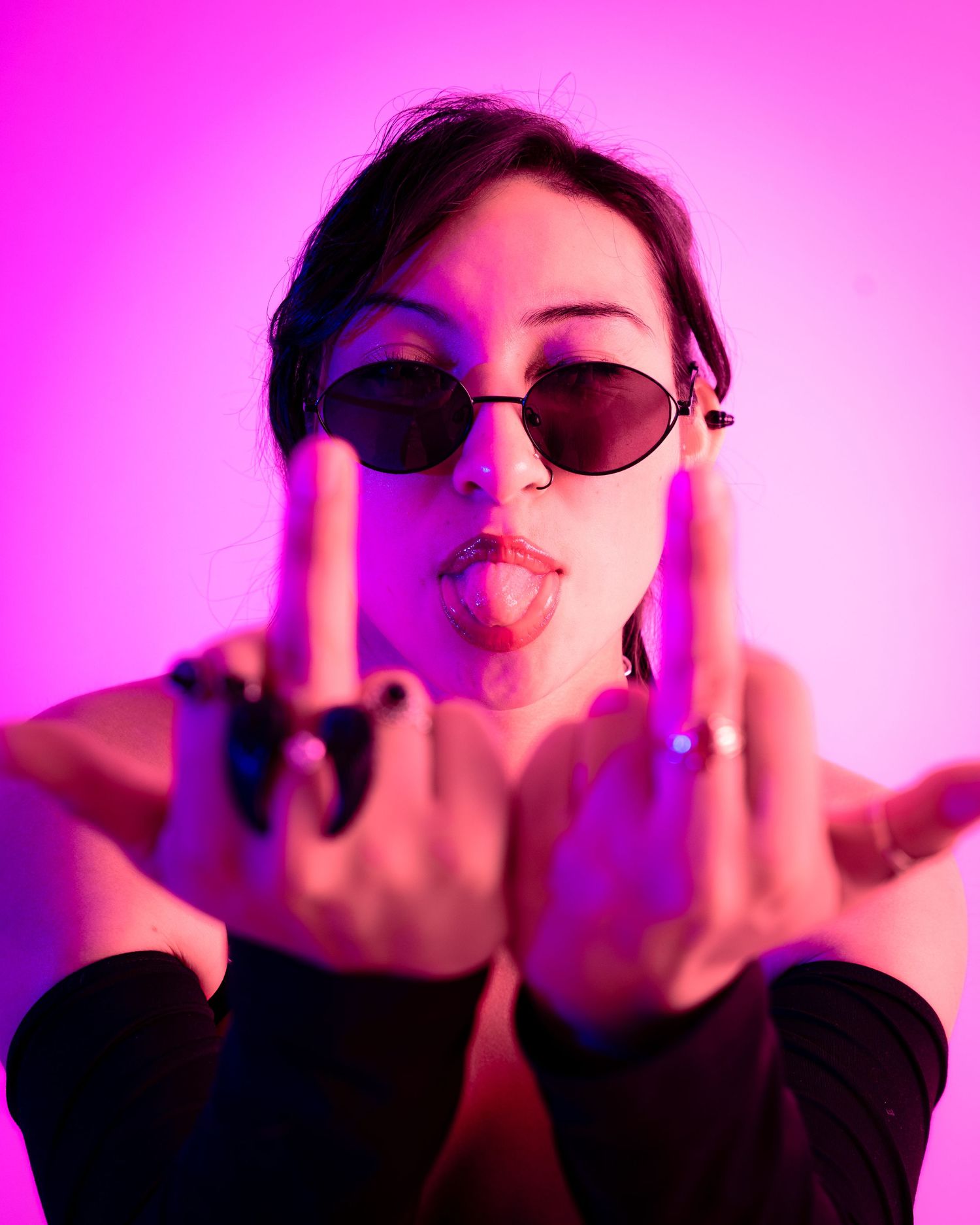 Edgy portrait with sunglasses making rebellious gesture against vibrant pink background.