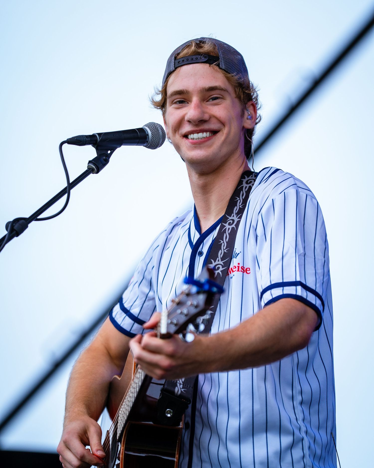 Performer in pinstriped jersey smiling while playing guitar during daytime outdoor performance.