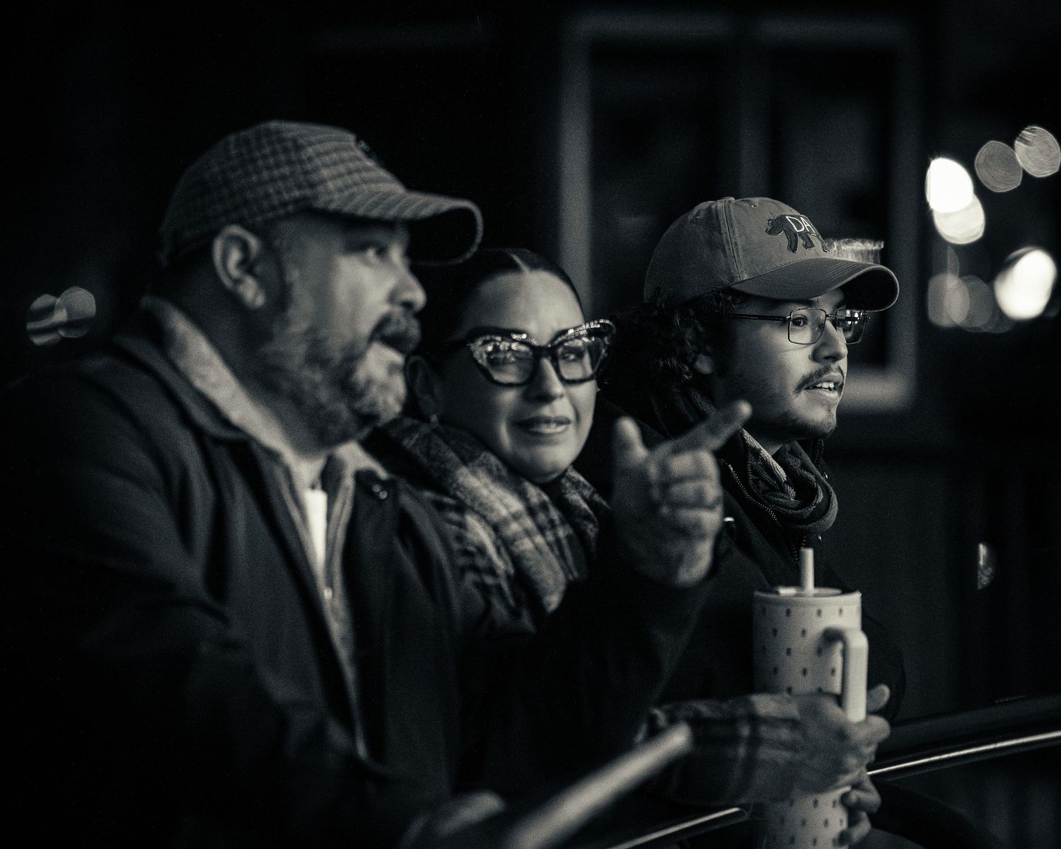 A black and white image shows spectators in baseball caps watching intently from the stands at night.