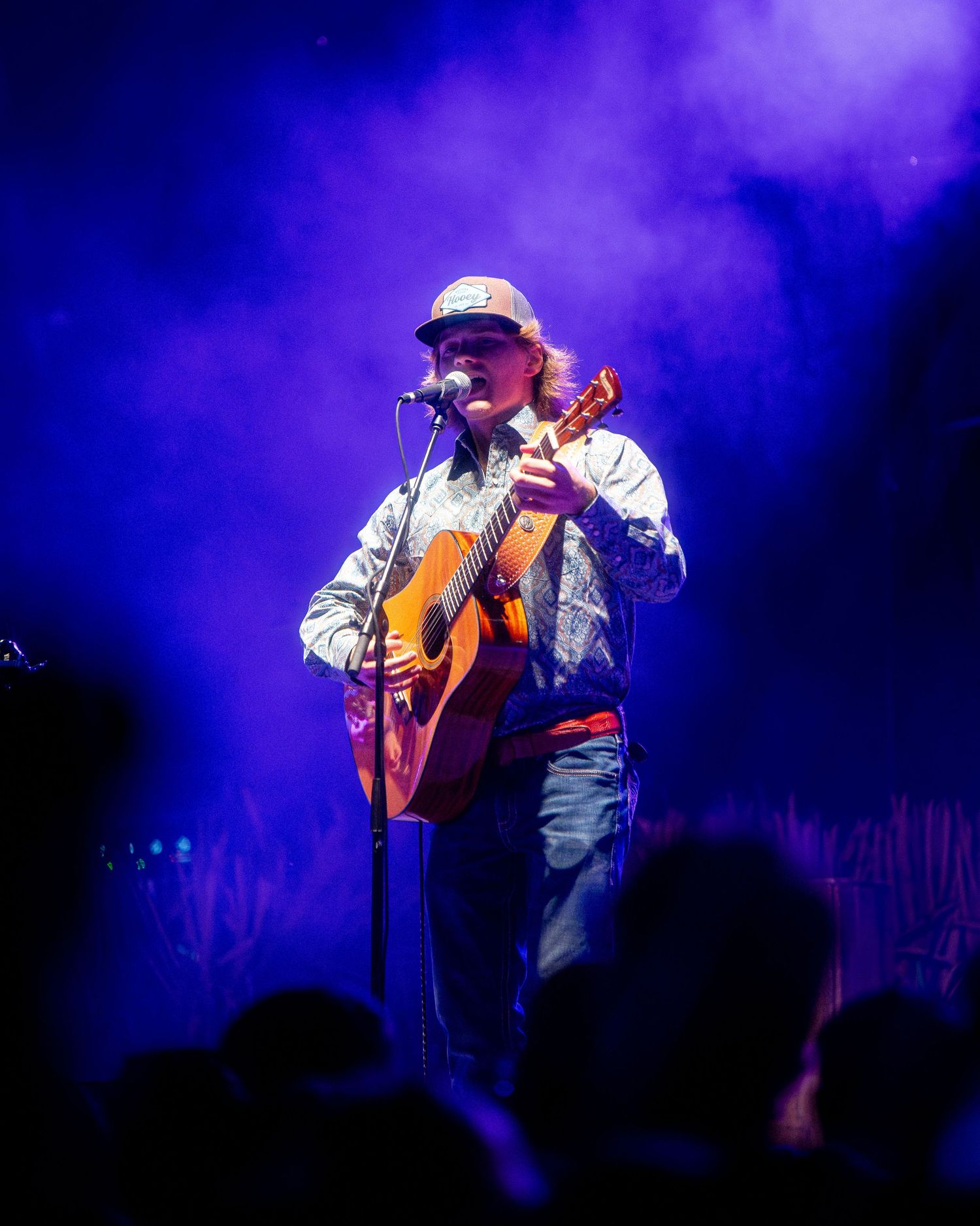Performer plays acoustic guitar on stage bathed in purple and blue atmospheric lighting.