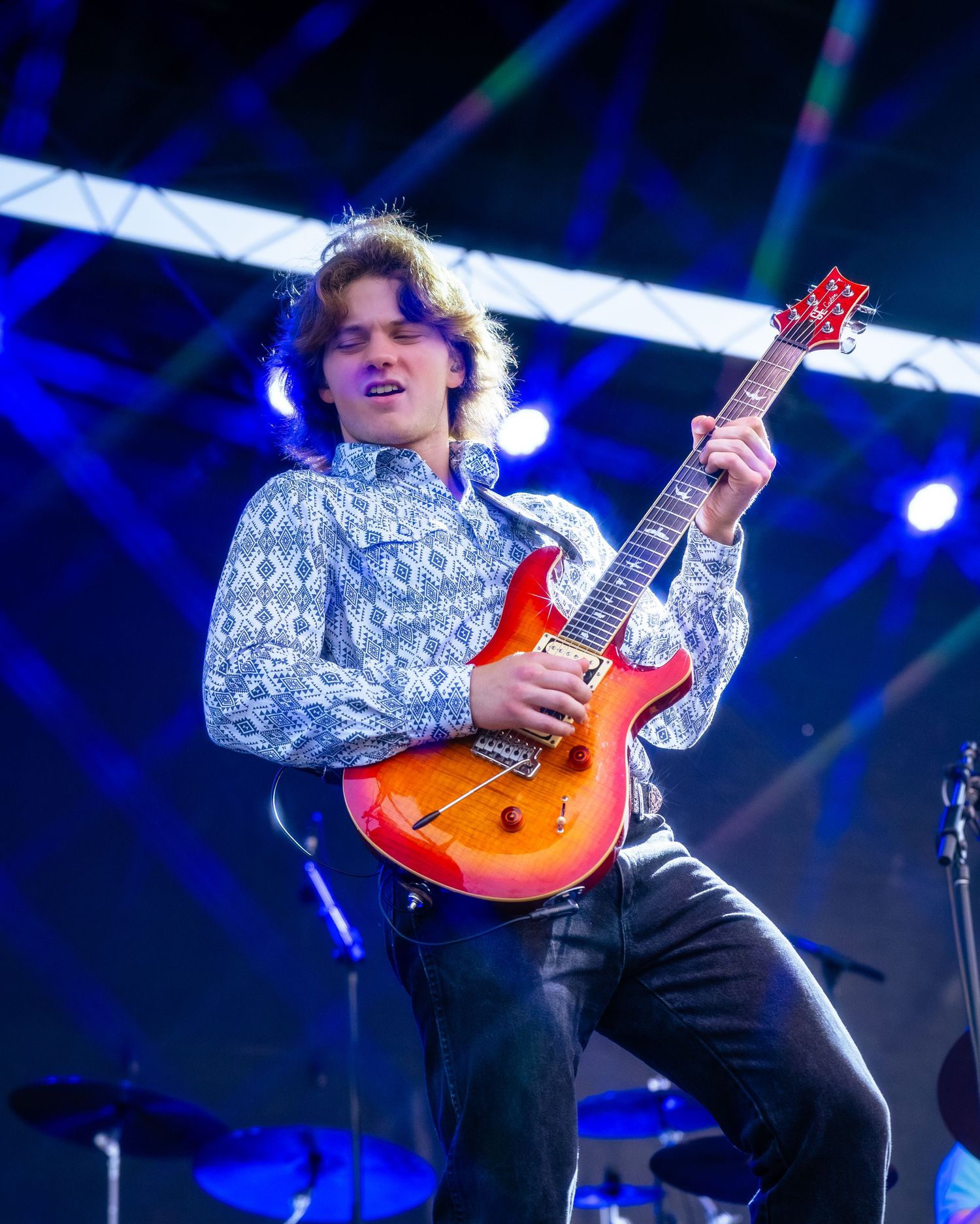 Performer playing sunburst guitar on stage with dramatic blue lighting and geometric backdrop.