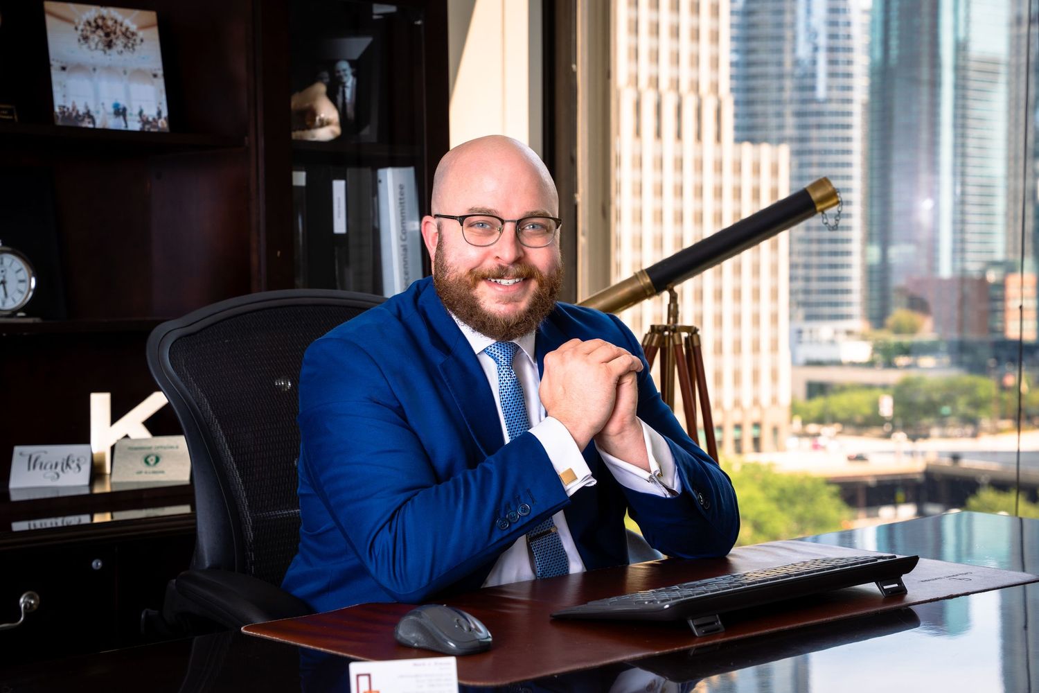 Executive in blue suit sits at desk with city skyline view through window.