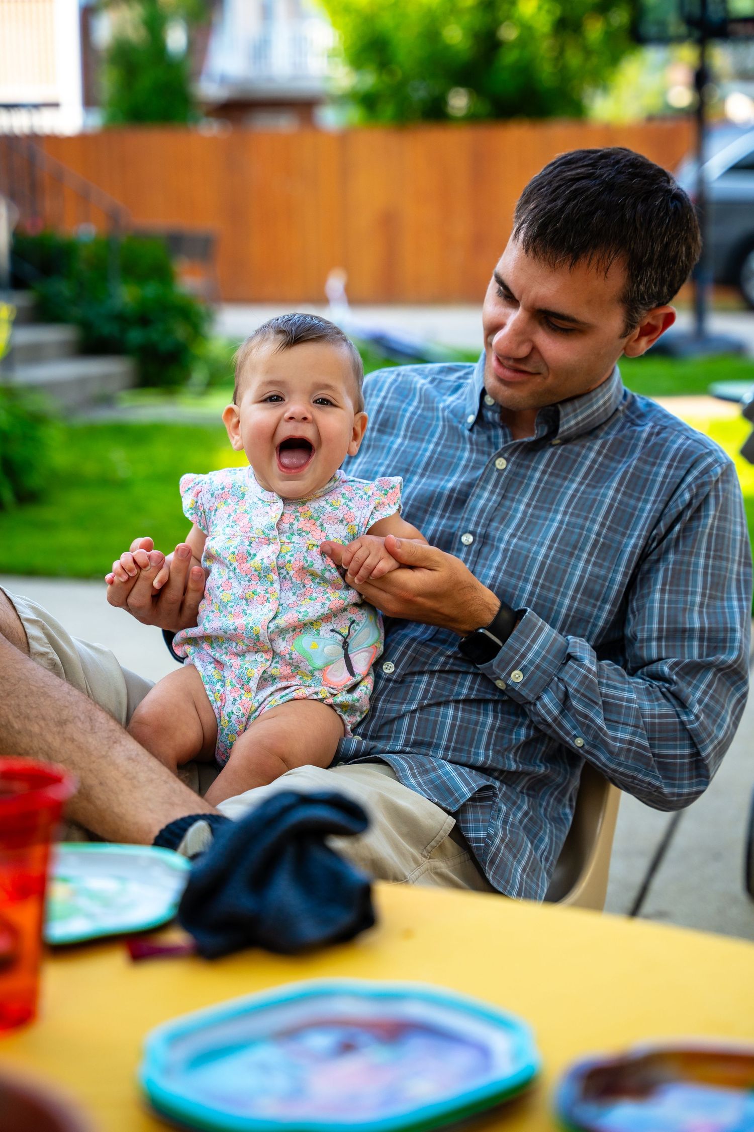 A baby laughs while being fed at an outdoor table with colorful dishes.