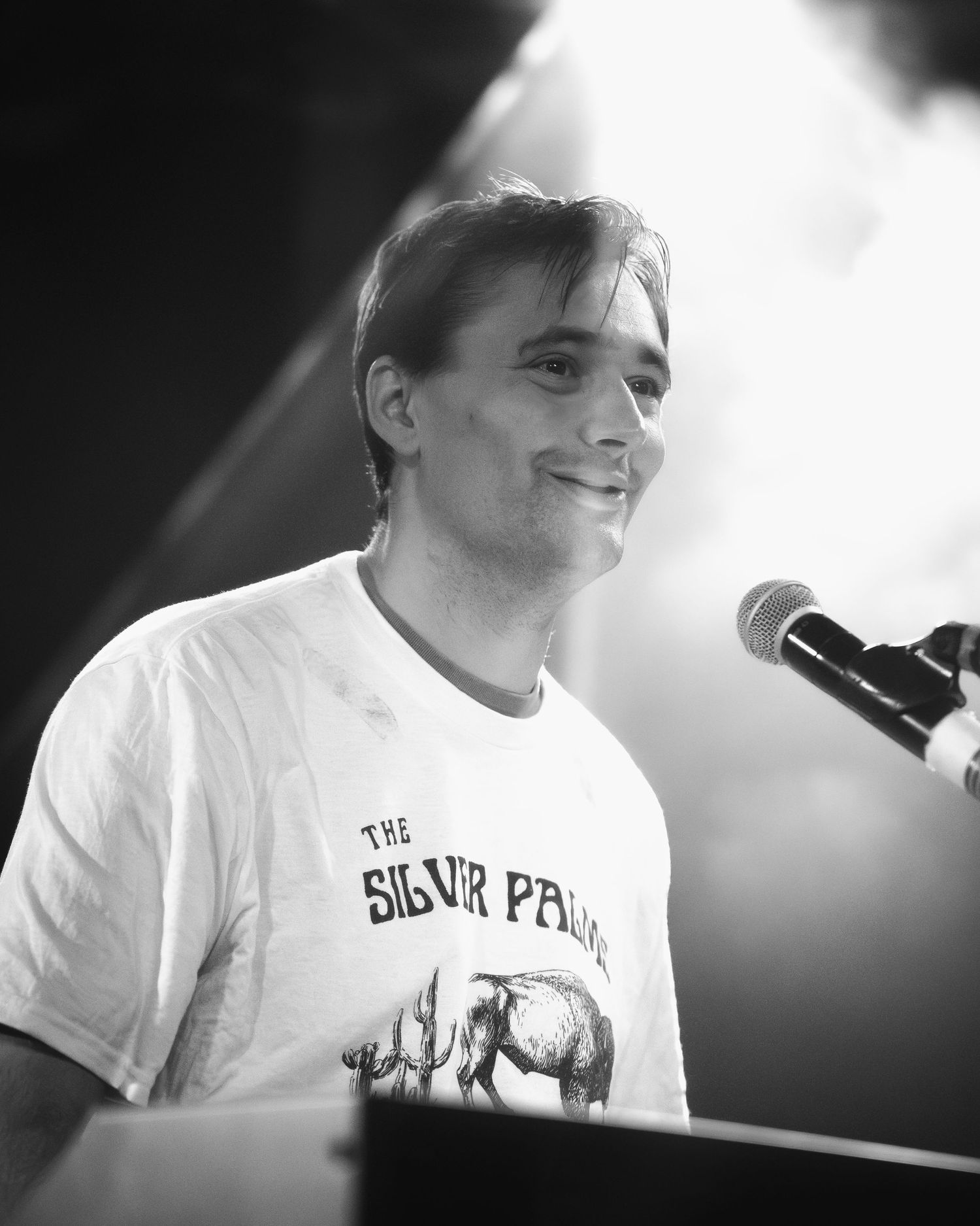 Concert performer in white t-shirt at microphone with bright stage lighting.