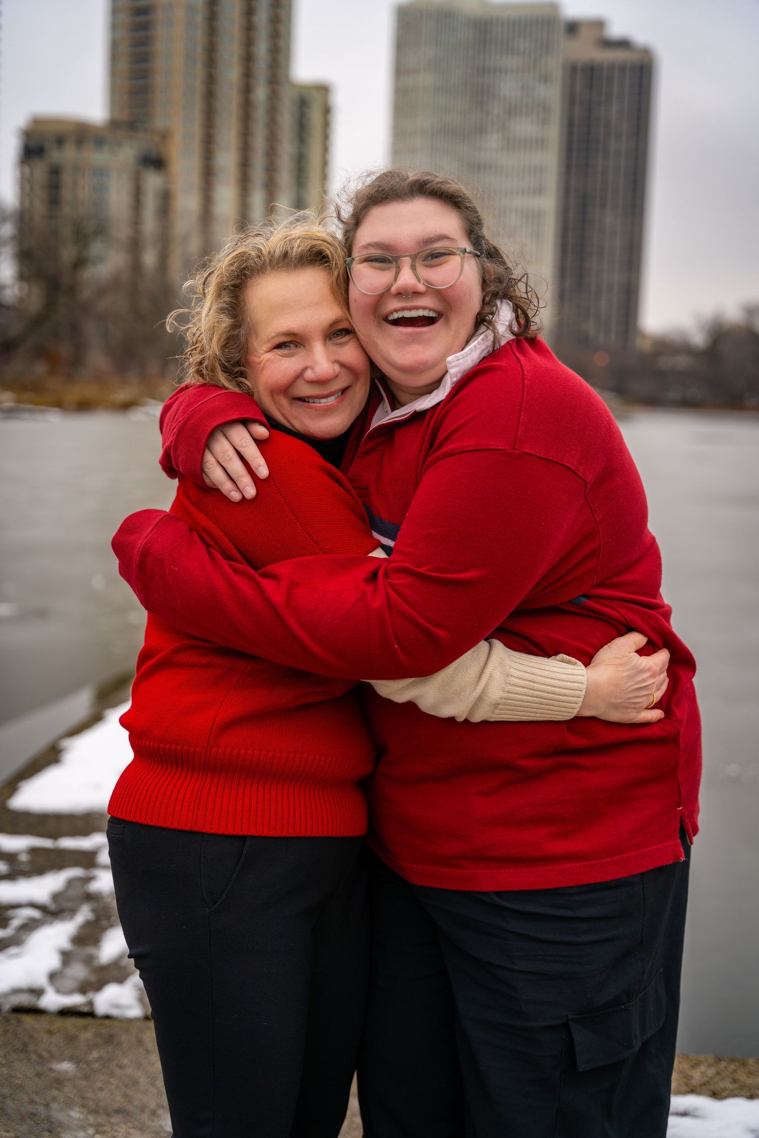 Two people in red sweaters share warm embraces outdoors on a winter day with city buildings in the background.