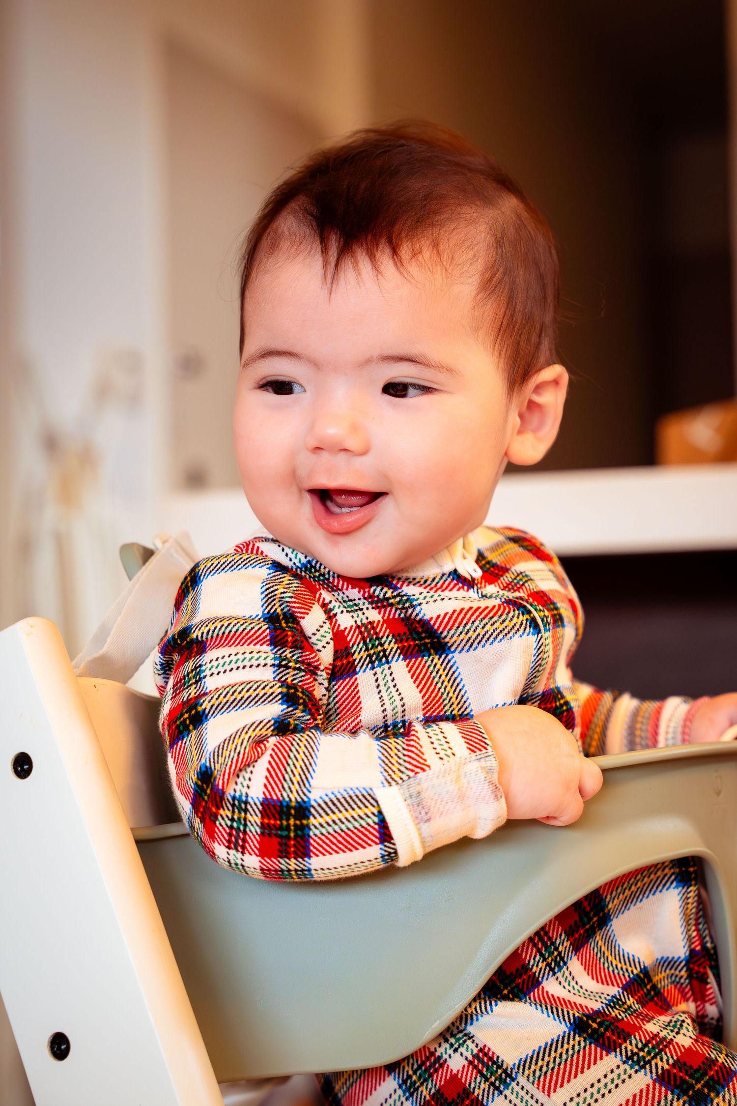 Happy baby wearing a red and white plaid shirt smiles while sitting in a high chair during mealtime.