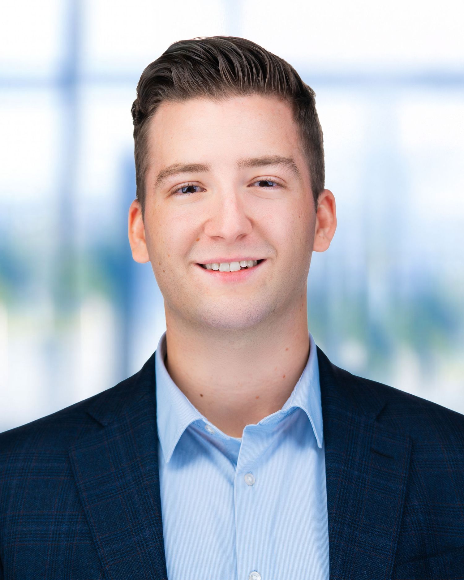 Corporate headshot of person wearing light blue shirt and navy blazer with bright background.