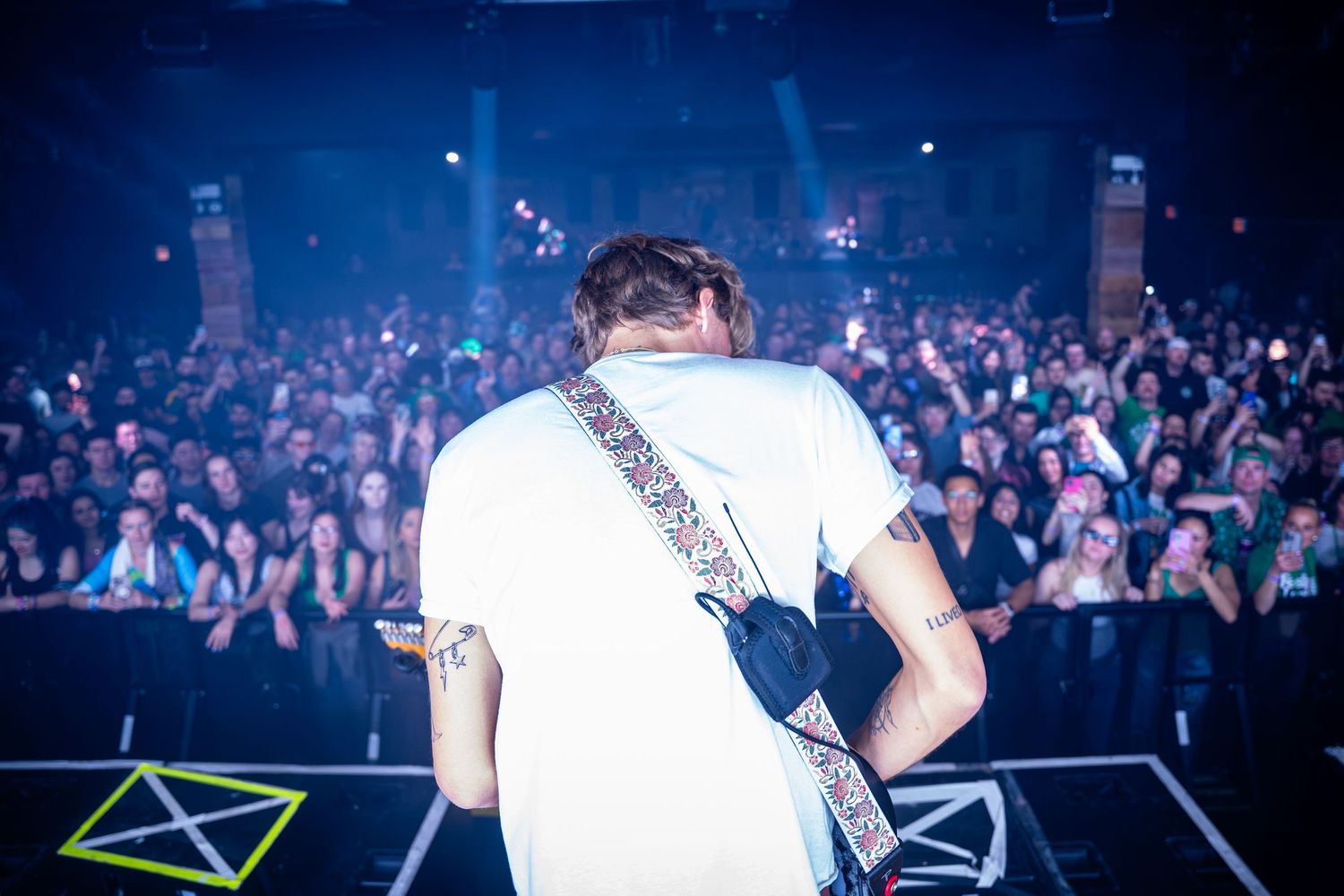 Concert performer facing large crowd from stage with blue lighting effects.