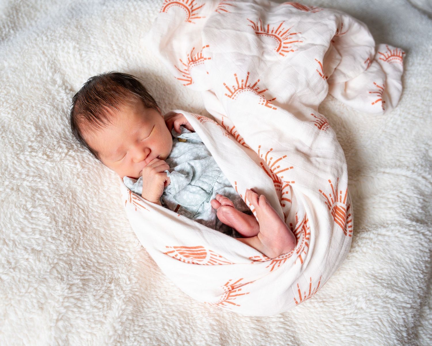 A newborn baby wearing white sleepwear rests peacefully on white textured bedding.