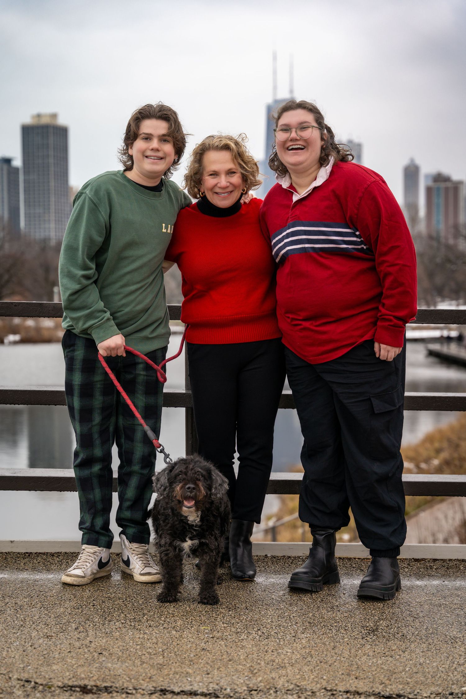Three people and a small brown dog pose together on a path with city buildings visible in the background.