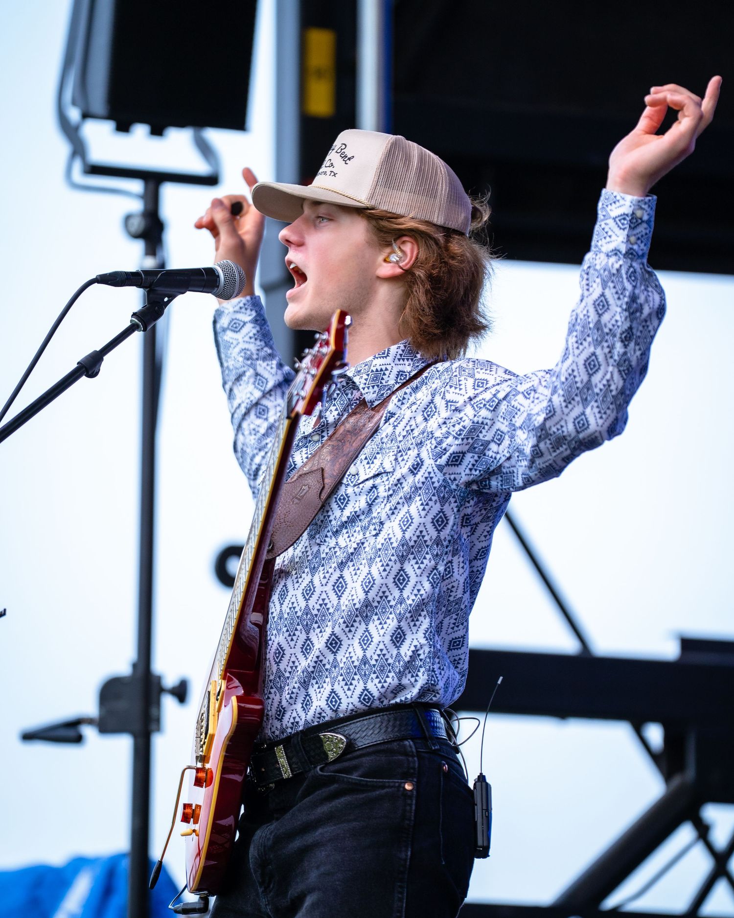 Musician in patterned shirt and cap performing energetically with raised arm on outdoor stage.