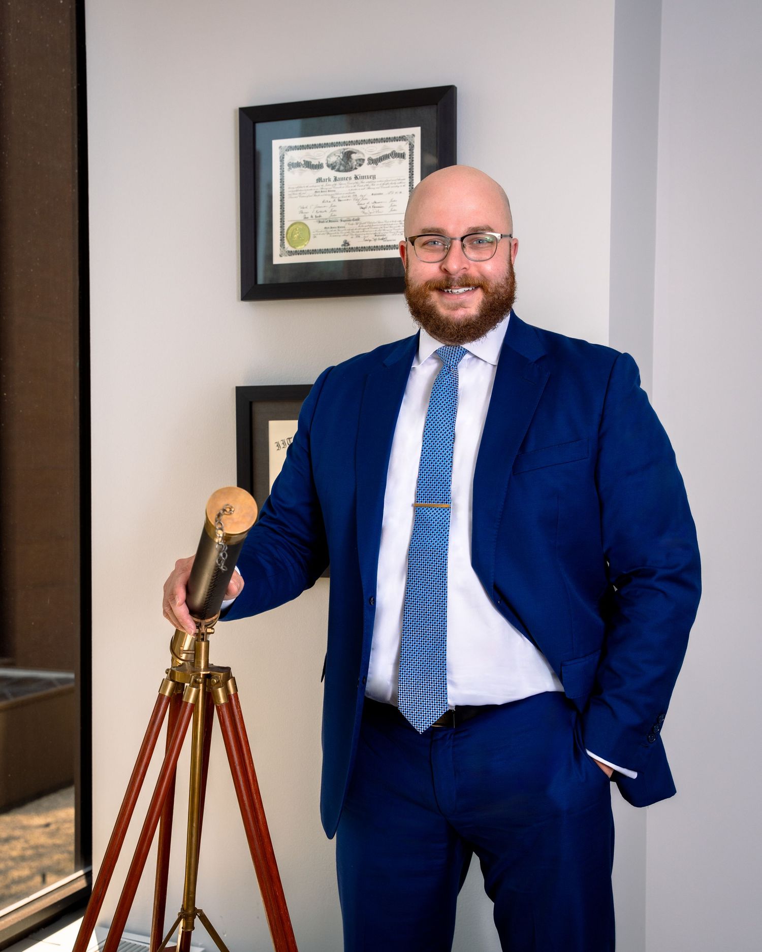 Professional in a navy blue suit poses with a vintage tripod in an office setting.