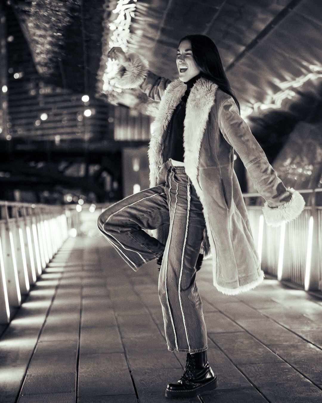 A person dances and spins on an illuminated walkway wearing a brown coat and jeans at night.